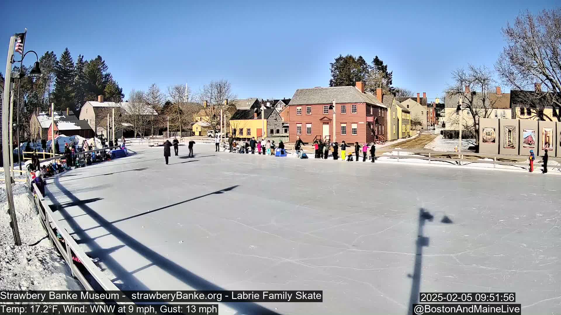 A group of people ice skate on an outdoor rink in front of several historic-looking buildings on a sunny day with a temperature of 17.2°F and a west-northwest wind.
