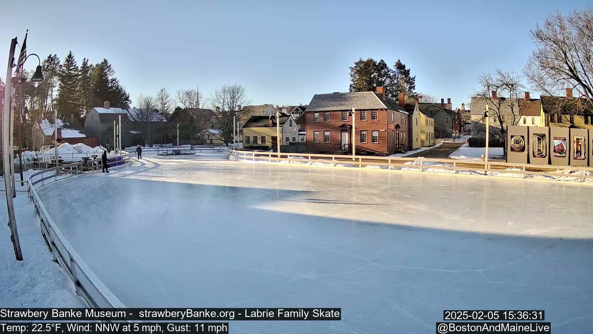 A mostly empty outdoor ice skating rink is situated in front of several historic-looking buildings on a sunny day with a northwest wind.