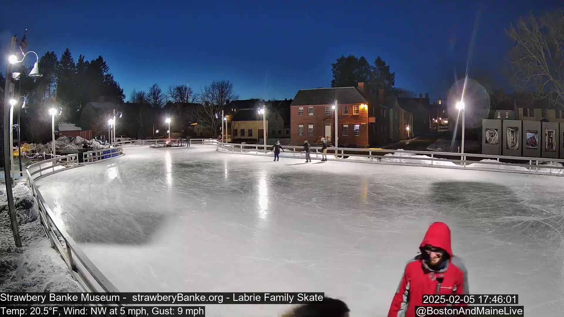 An outdoor ice skating rink at night, illuminated by lights, with several people ice skating and a person in a red hooded jacket walking toward the camera under a clear, dark blue twilight sky.