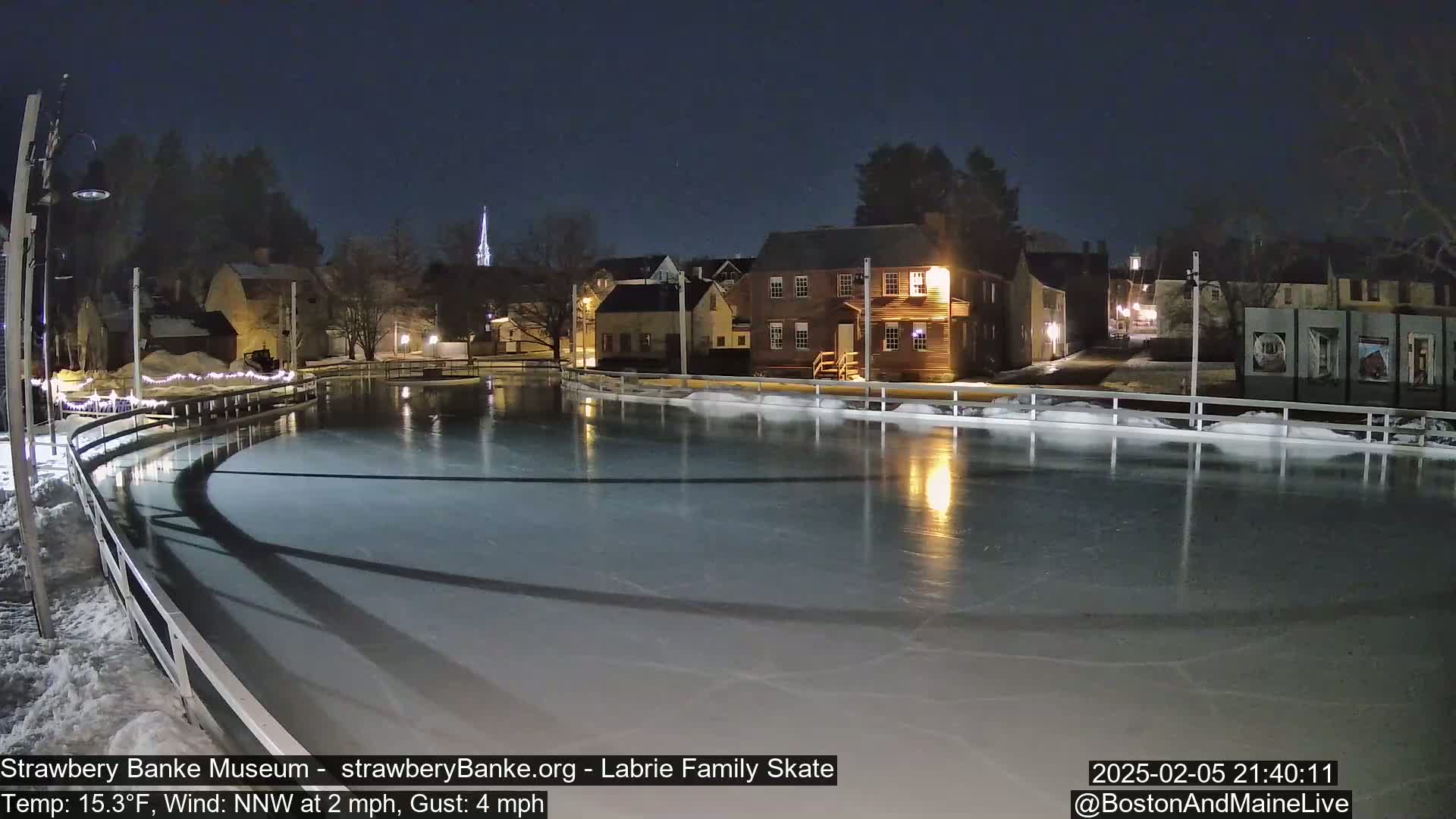 A nighttime view of an outdoor ice skating rink, illuminated, with buildings and trees in the background under a clear, dark sky.