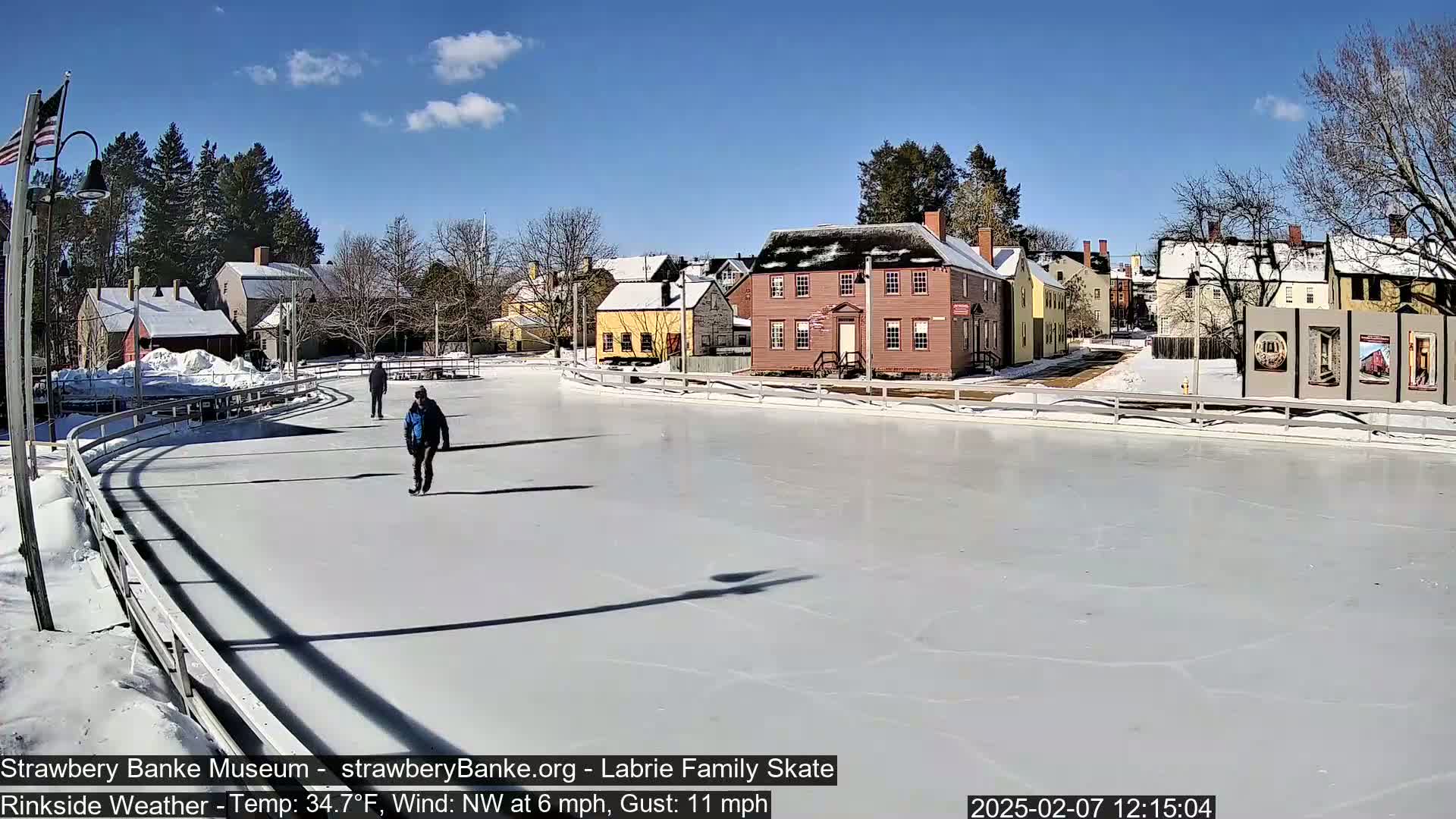 Two people ice skate on a rink in front of a row of historic buildings on a sunny day with a northwest wind.