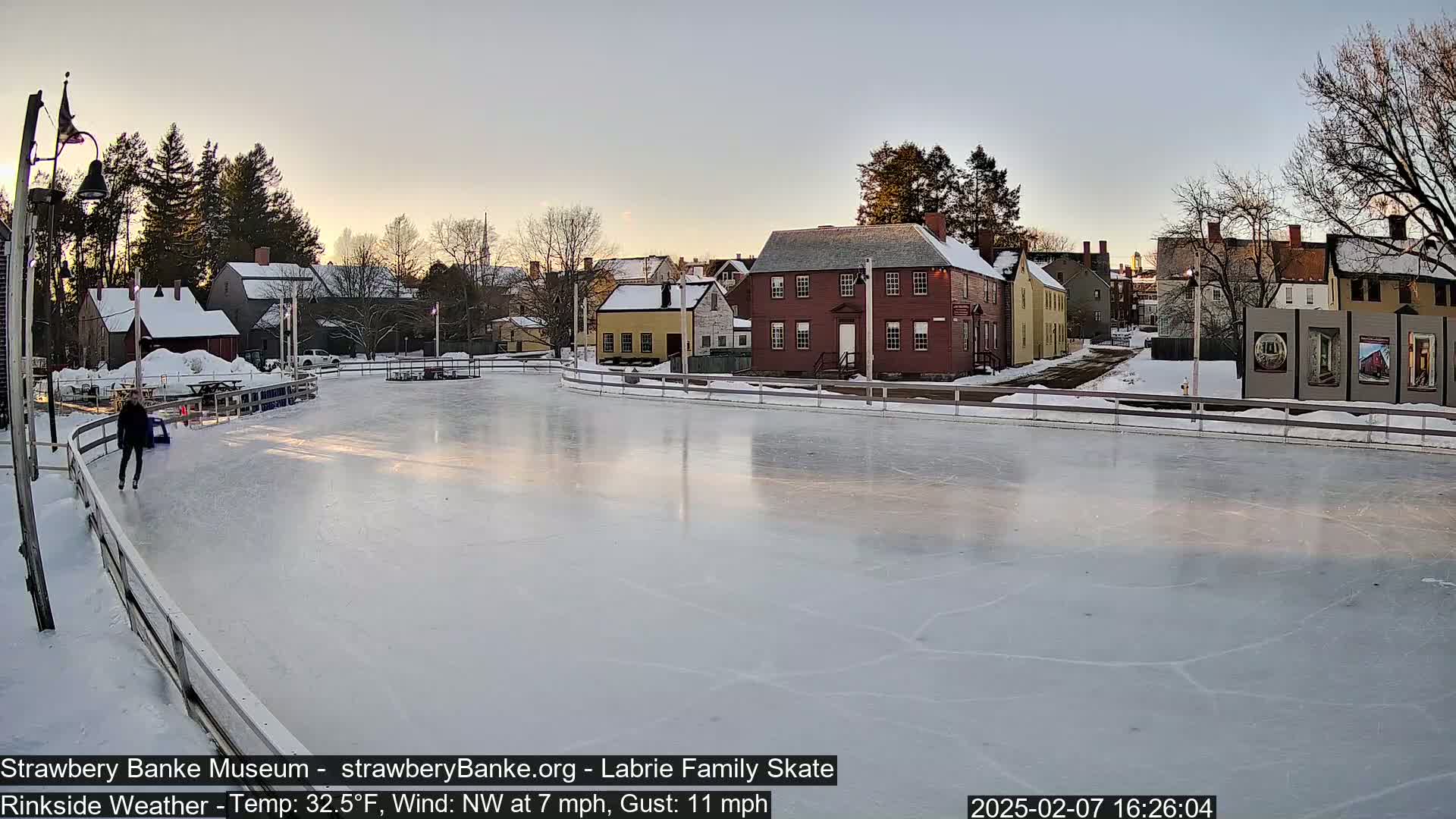 A person ice skates on a rink surrounded by snow-covered colonial-style buildings under a mostly clear, twilight sky.