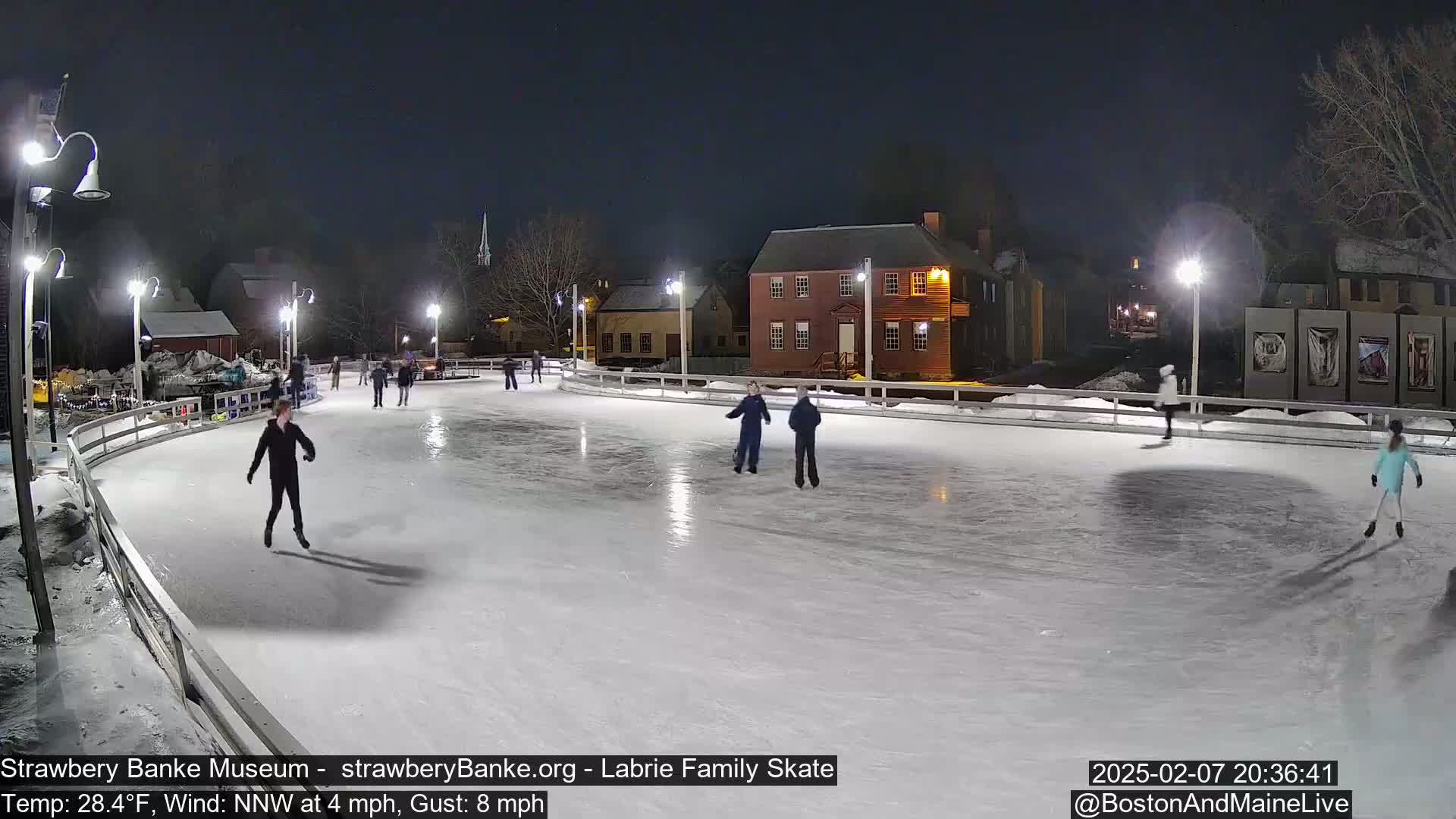 Several people ice skate at night on an outdoor rink illuminated by lights, with a cold wind blowing.