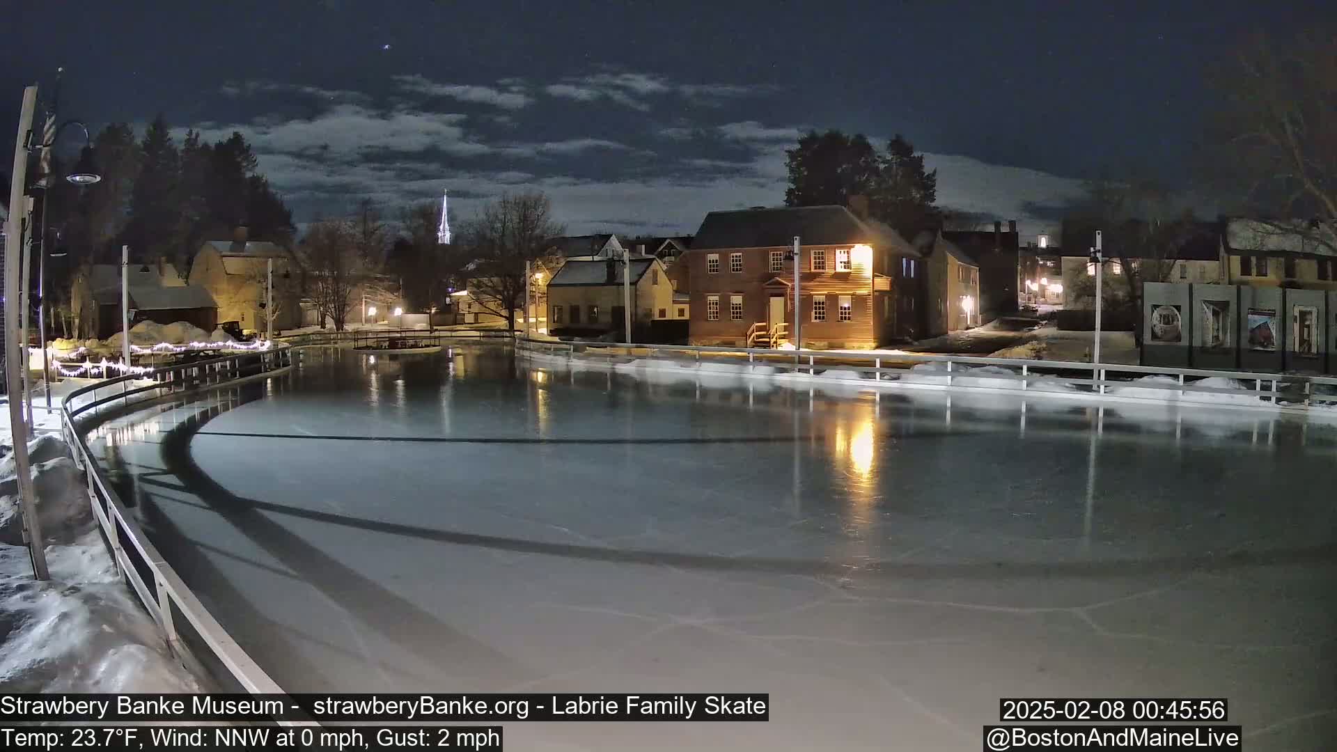 A nighttime view of an ice skating rink illuminated by lights, surrounded by snow-covered buildings and trees under a partly cloudy sky.