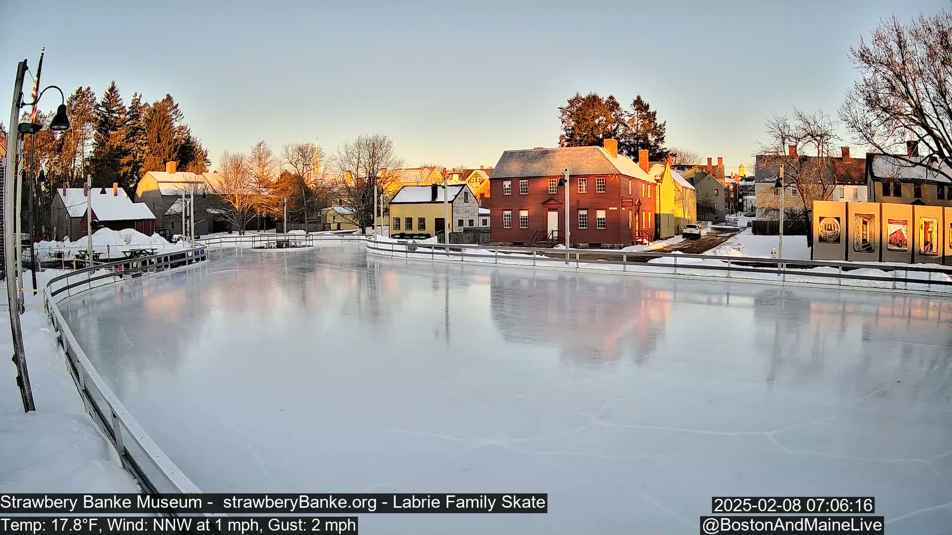 A frozen skating rink reflects the sunset light on its surface, situated beside several historic-looking buildings in a snowy, winter scene.