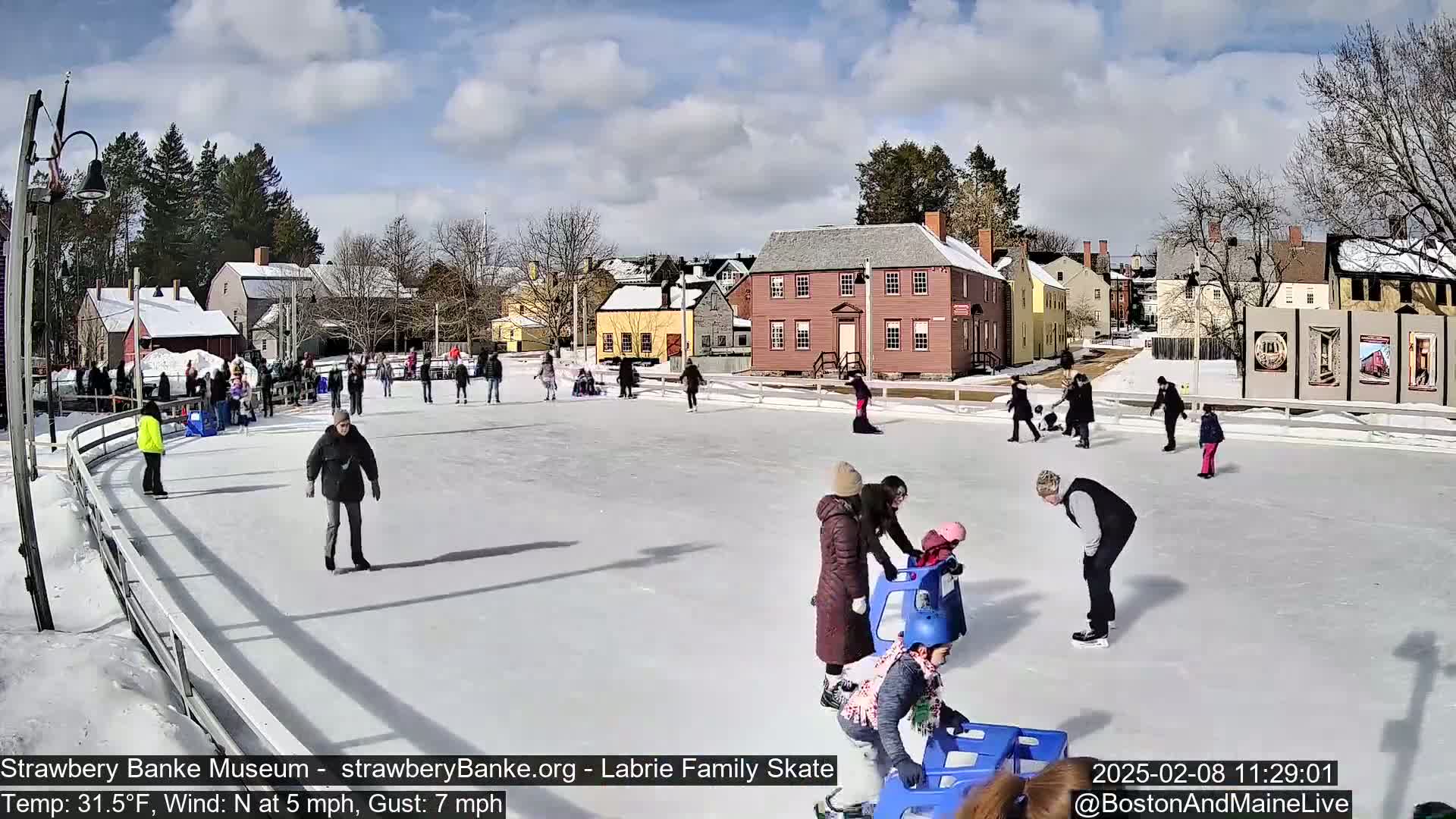 People ice skate on an outdoor rink in front of historic buildings under a partly sunny sky.