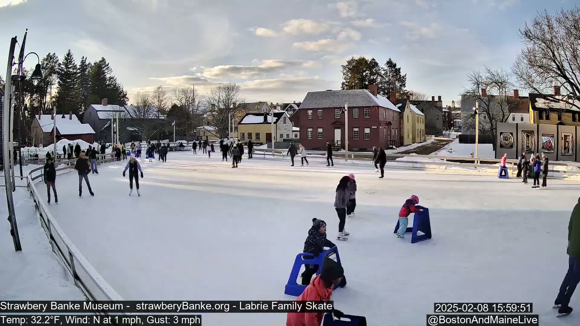 Many people ice skate on an outdoor rink, surrounded by snow-covered buildings under a partly cloudy sky.