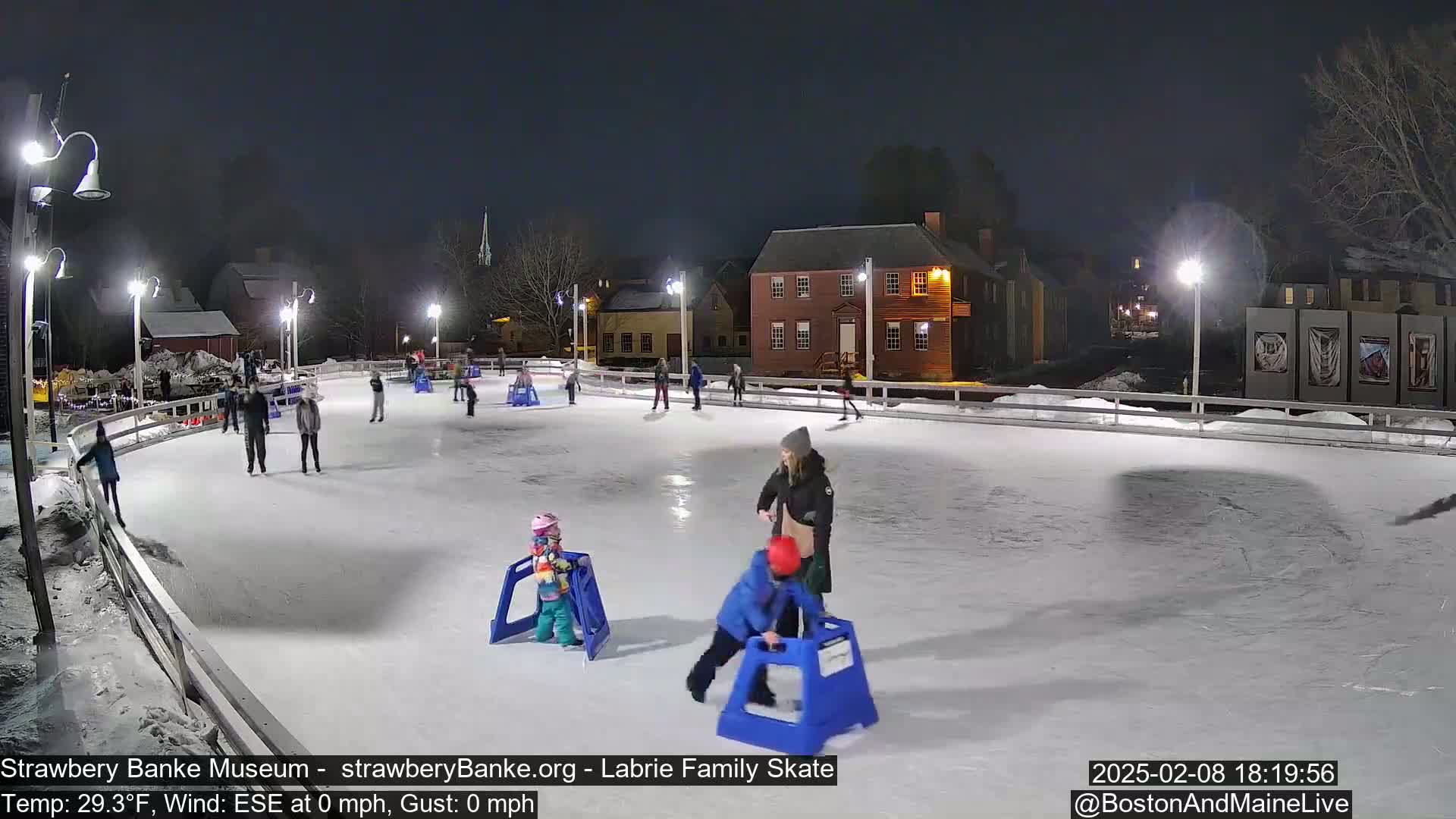 At night, several people ice skate on an outdoor rink illuminated by lights, with a calm wind and a temperature of 29.3°F.