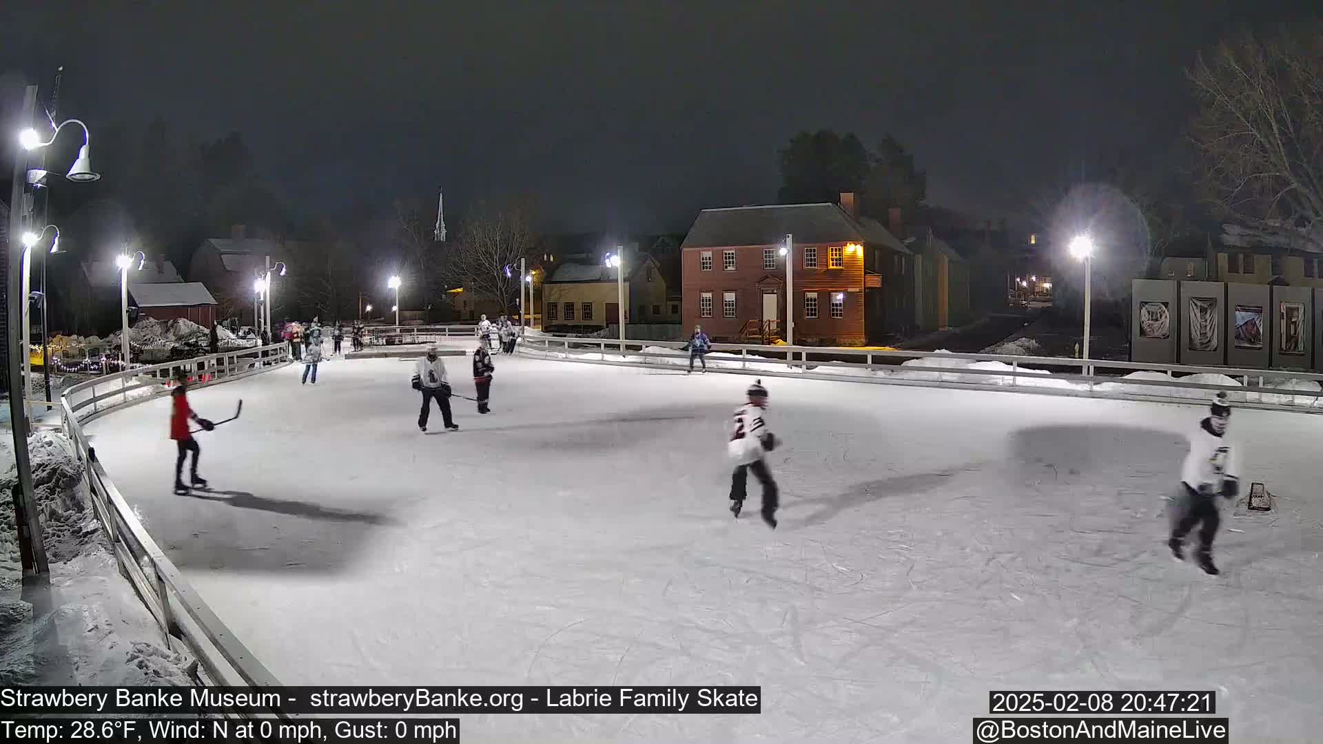 Several people are ice skating and playing hockey on an outdoor rink at night under bright lights.