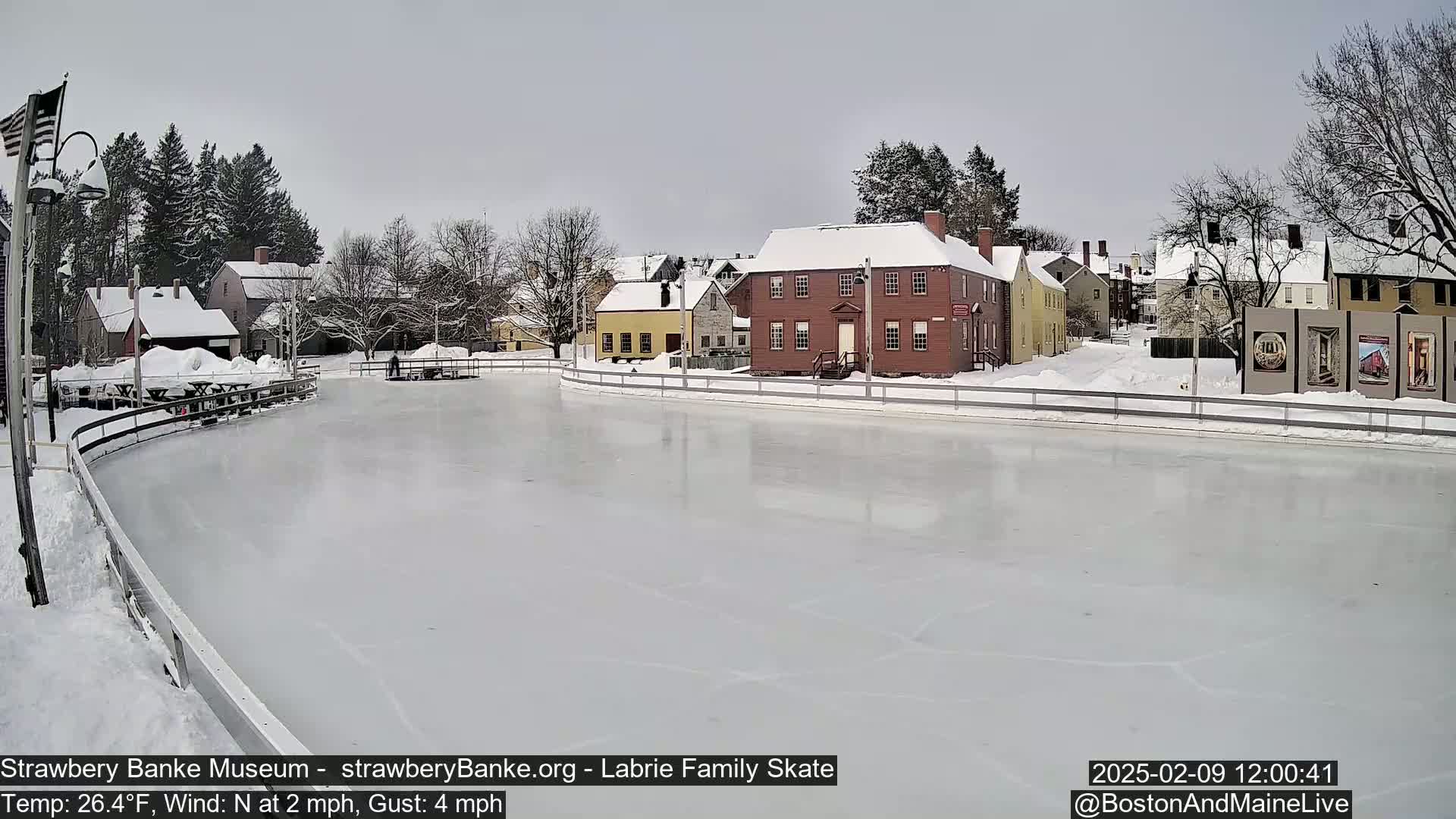 A snow-covered outdoor ice skating rink sits in front of several historic-looking buildings under a cloudy sky.