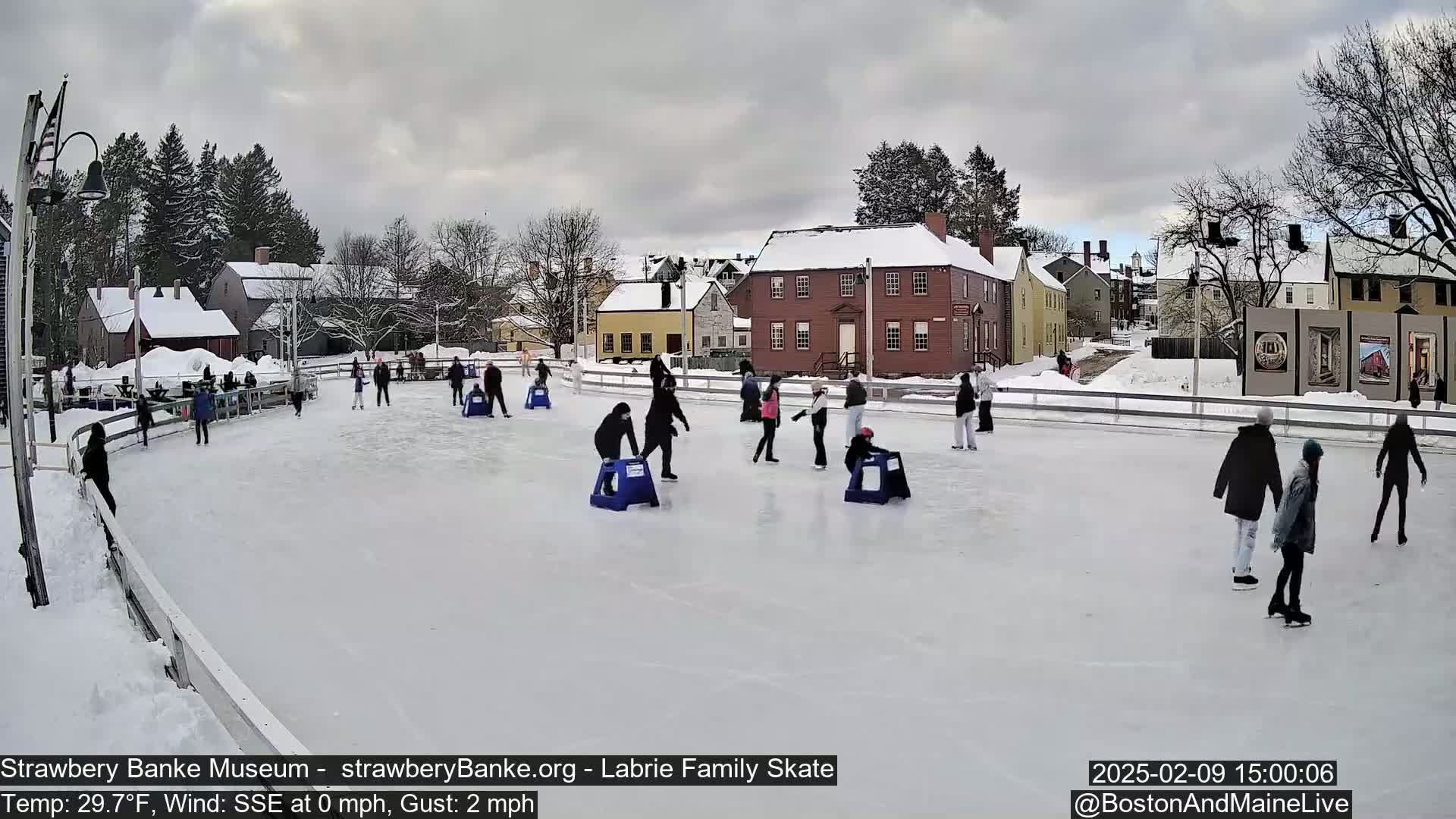People ice skate on an outdoor rink in front of snow-covered buildings under an overcast sky.