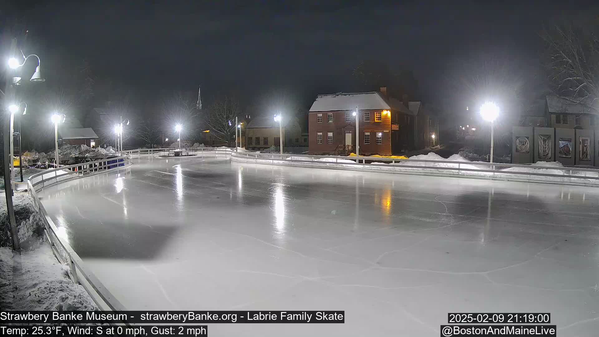 A brightly lit outdoor ice skating rink at night, surrounded by snow-covered buildings and trees under a dark sky.