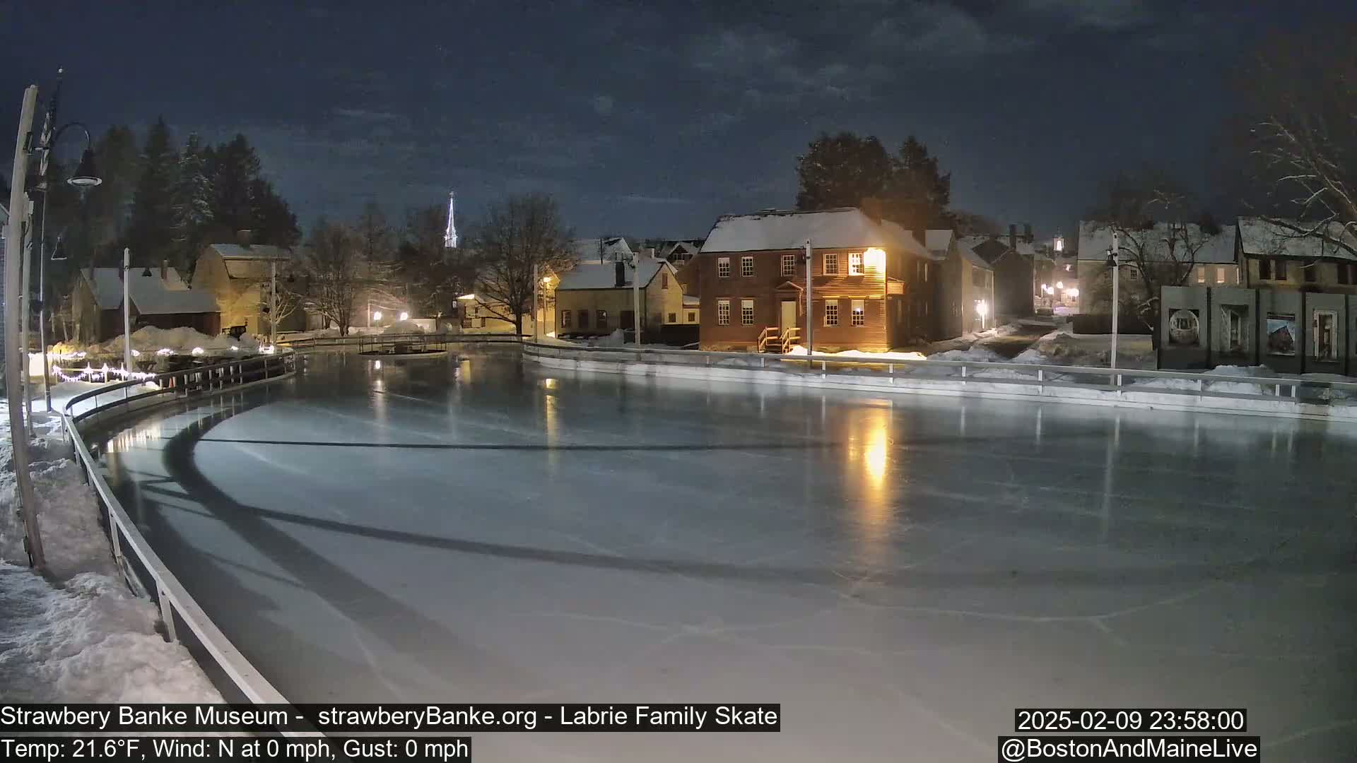 A nighttime view of an ice skating rink illuminated by lights, surrounded by snow-covered buildings under a mostly clear night sky.