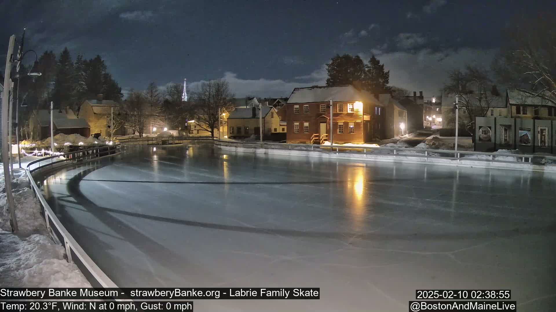 A nighttime view of an ice skating rink, illuminated by lights, situated beside snow-covered buildings under a partly cloudy night sky.