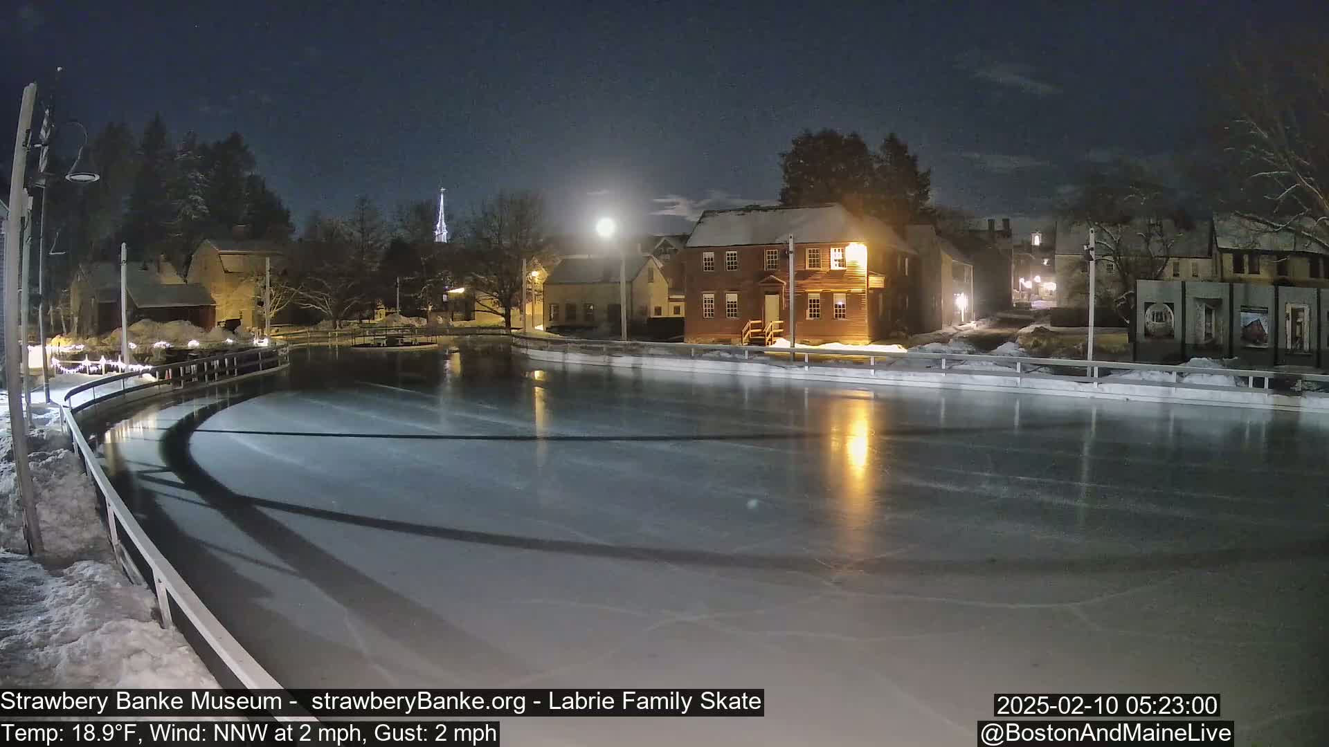 A nighttime view of a frozen skating rink illuminated by lights, surrounded by snow-covered buildings under a mostly clear sky with a moon visible.
