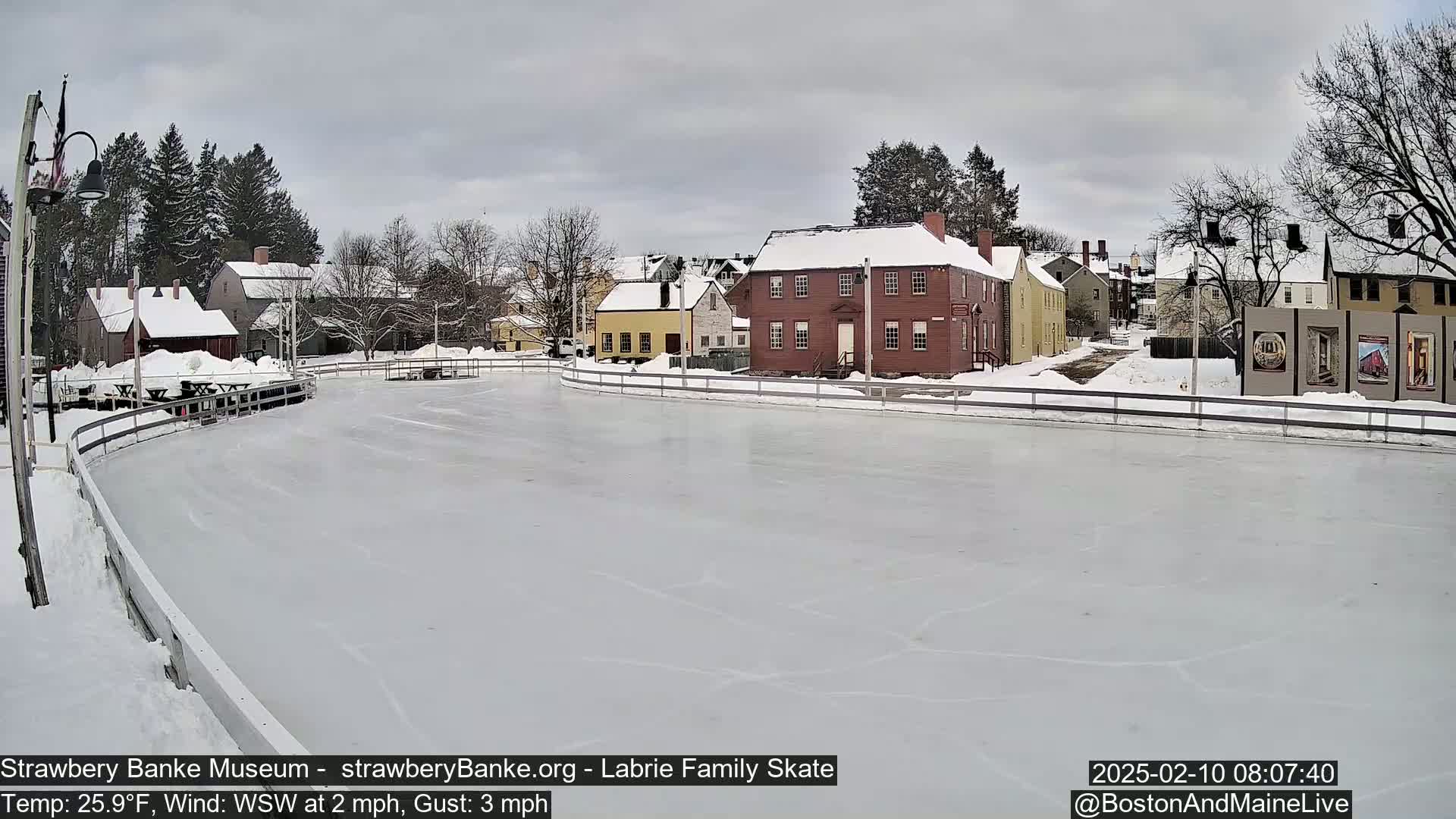 A snow-covered outdoor ice skating rink is situated in front of several historic-looking buildings under an overcast sky.