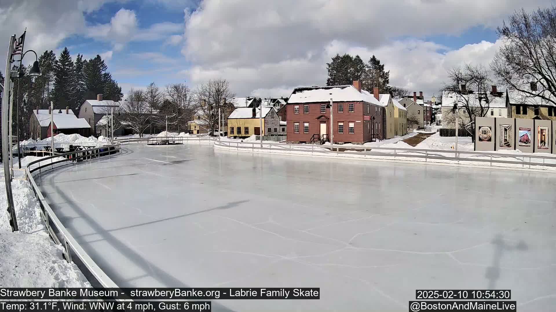 A snow-covered outdoor ice rink sits before a row of historic-looking buildings under a partly cloudy sky.