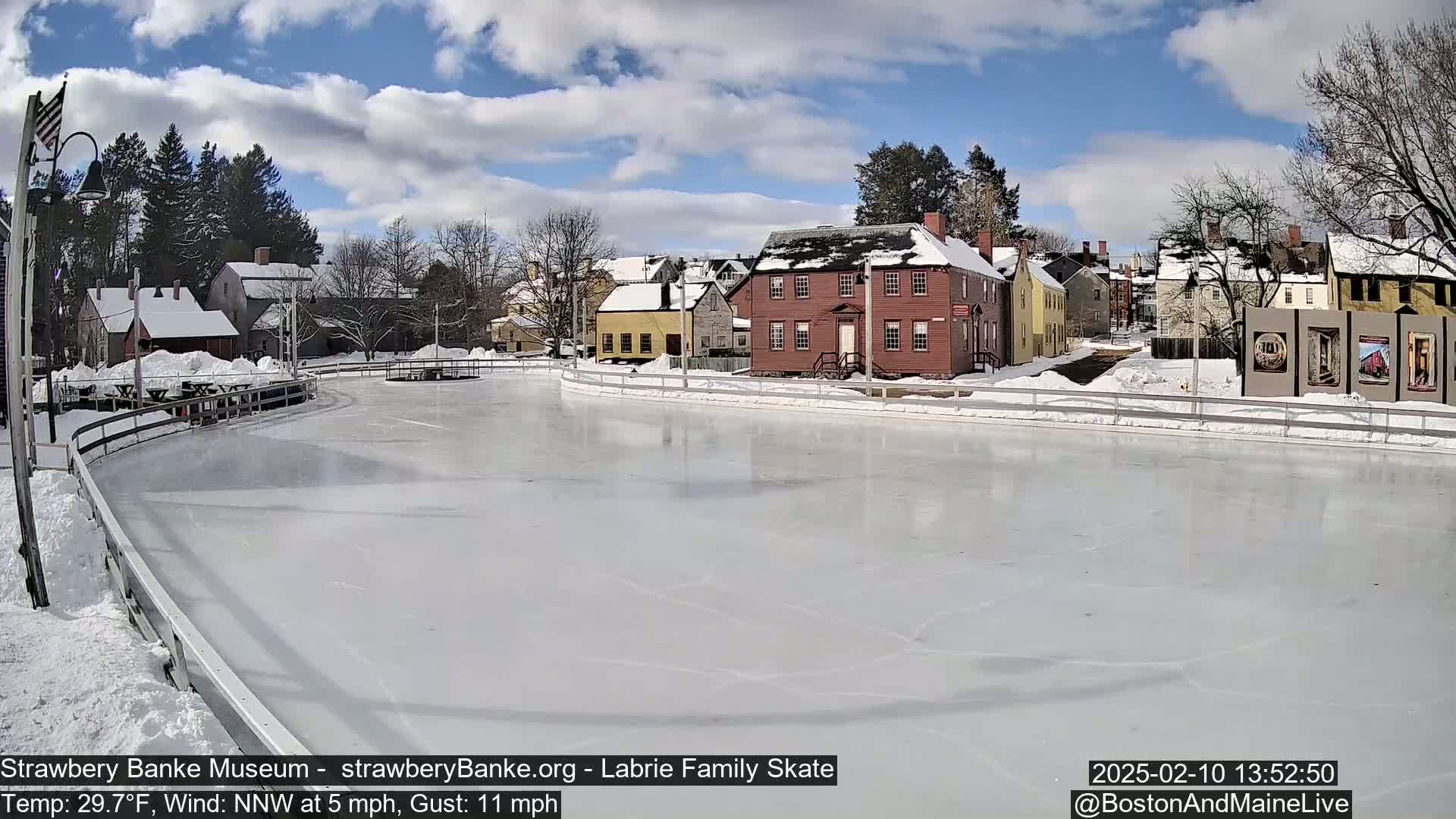 A frozen skating rink is situated in front of a row of historic-looking buildings on a partly cloudy winter day with a light wind.