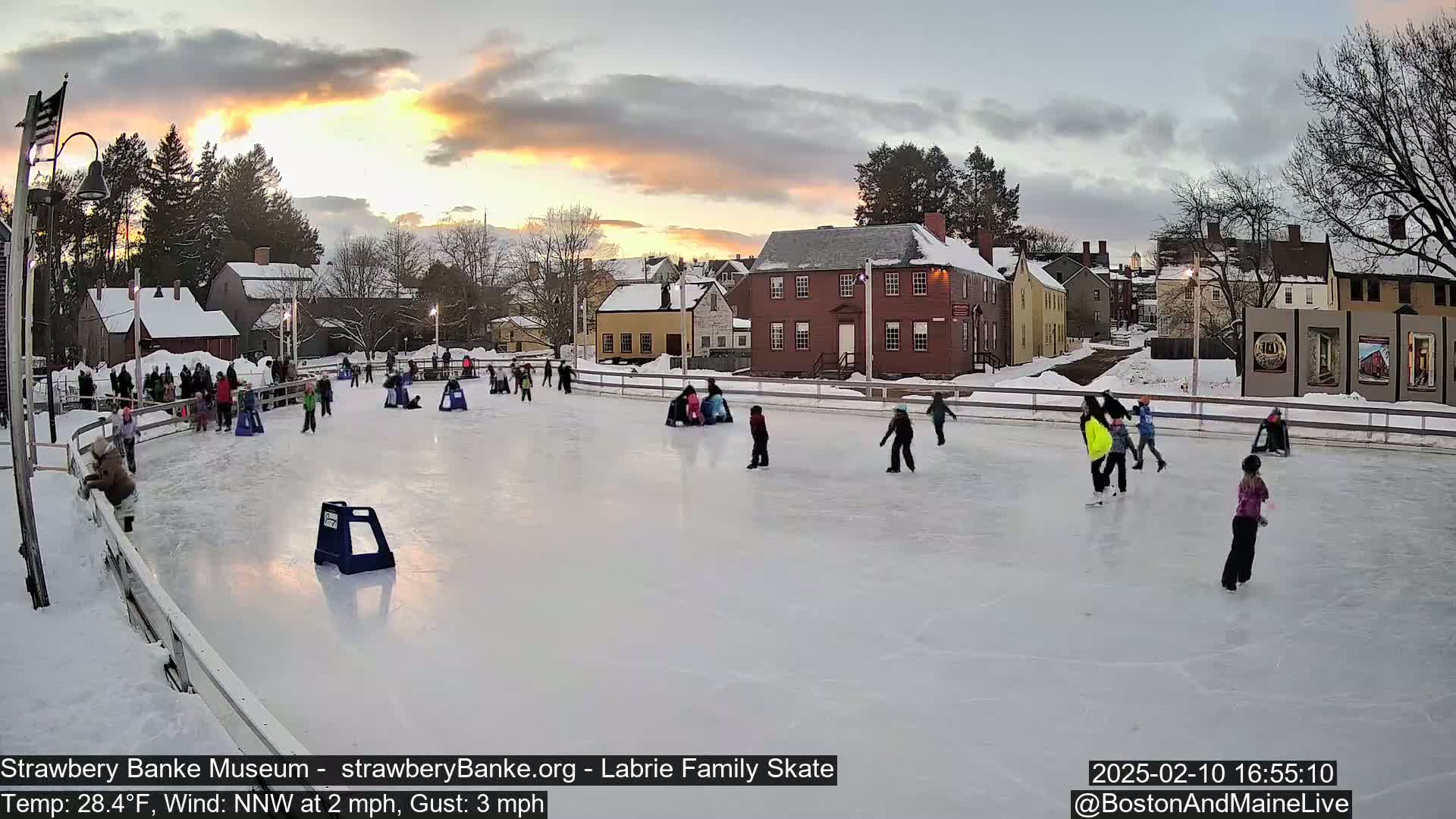 An outdoor ice skating rink, surrounded by historic buildings, is bustling with skaters during a sunset with a temperature of 28.4°F, a northwest wind of 2 mph, and gusts up to 3 mph.