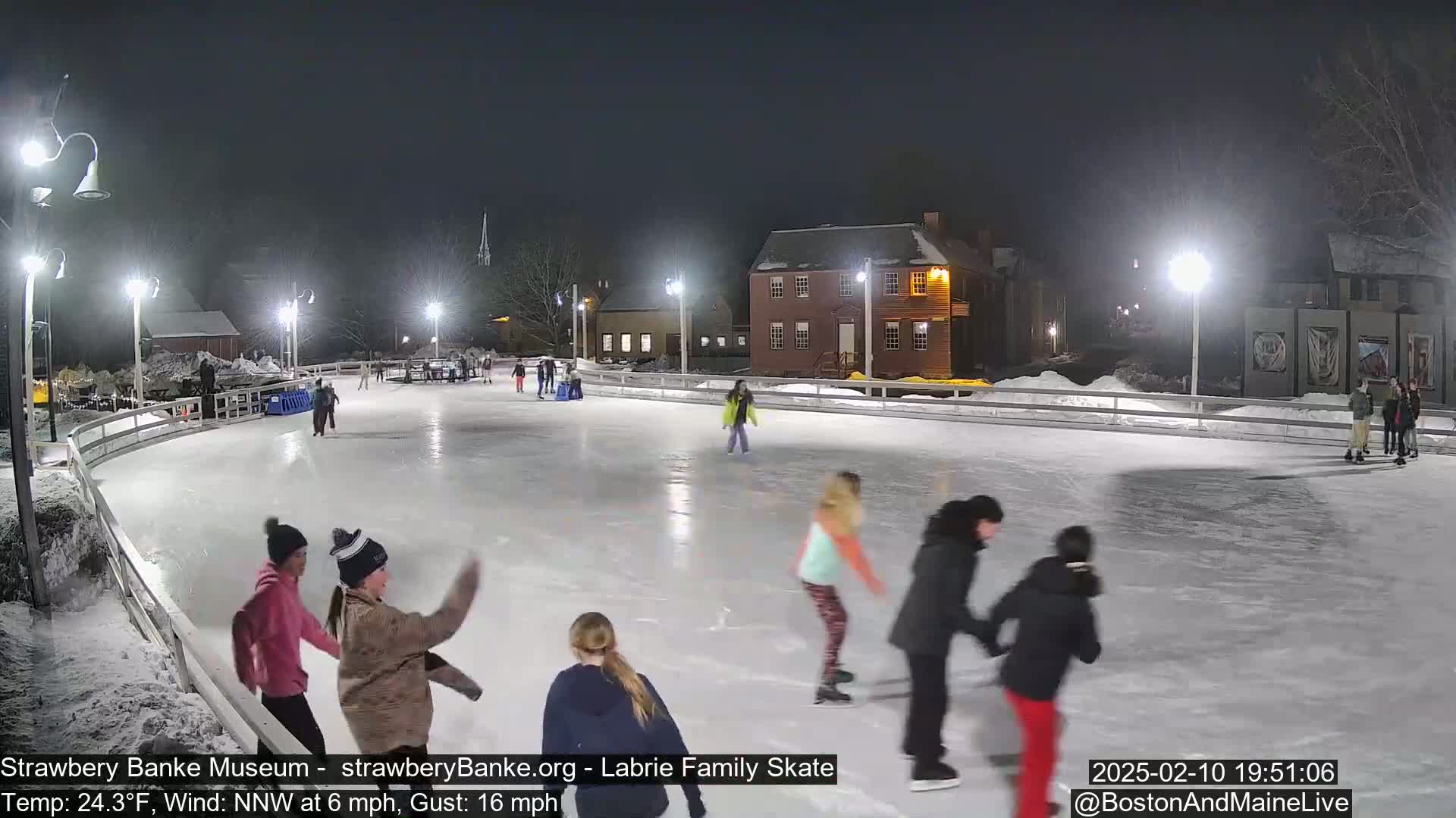 Several people ice skate on an outdoor rink at night, under bright lights, with a light snow cover on the ground.