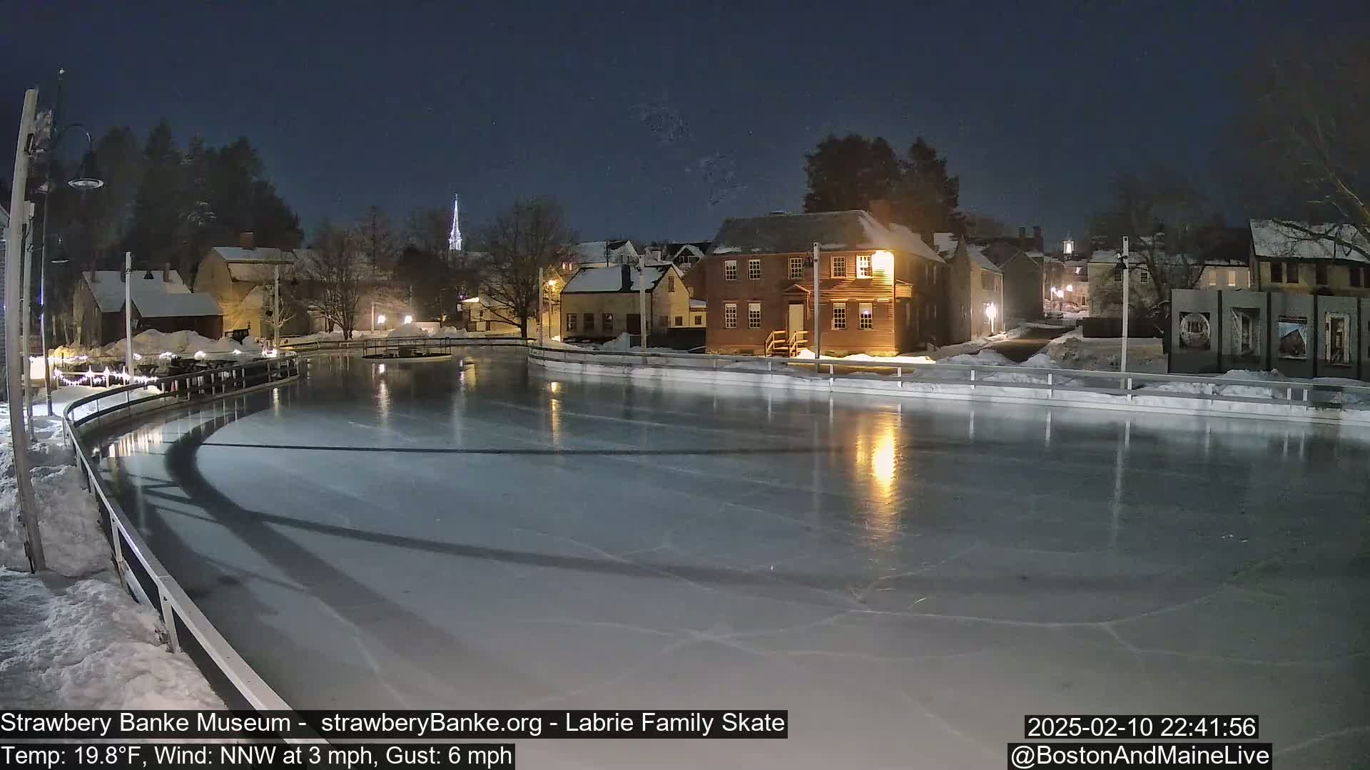 A nighttime view of an ice skating rink, illuminated by surrounding buildings, under a clear, dark sky.