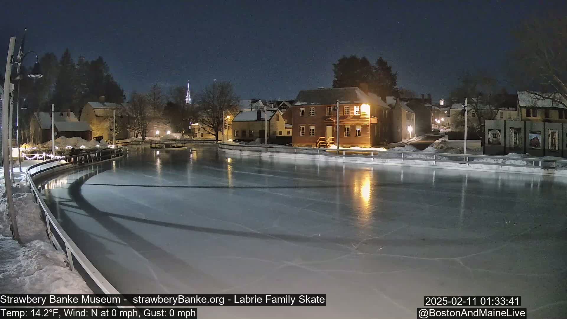 A nighttime view of an outdoor ice skating rink in a snow-covered village with calm, clear skies.