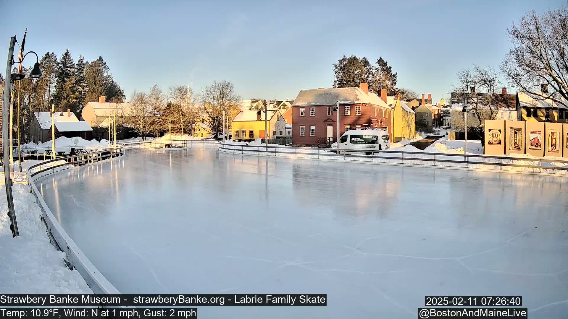 A mostly empty outdoor ice skating rink sits in front of historic-looking buildings on a sunny, cold winter day.