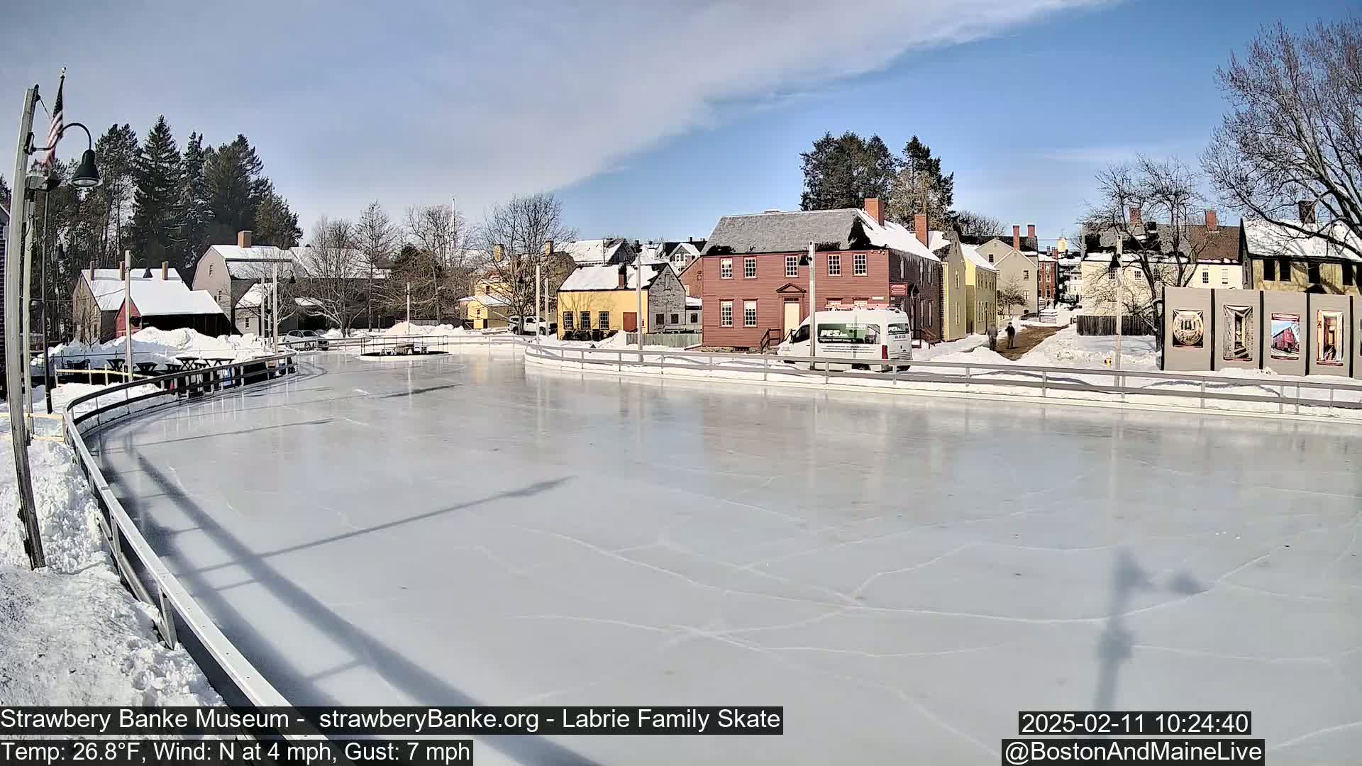 A sunny, partially cloudy day reveals an outdoor ice skating rink surrounded by historic-looking buildings and snow-covered ground.