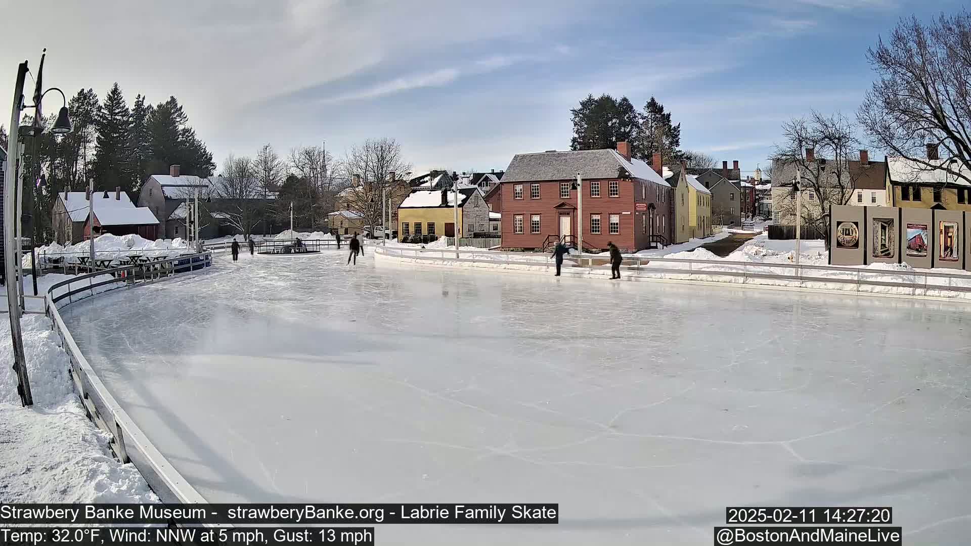 A mostly sunny day shows several people ice skating on an outdoor rink surrounded by historic-looking buildings and snow-covered ground.