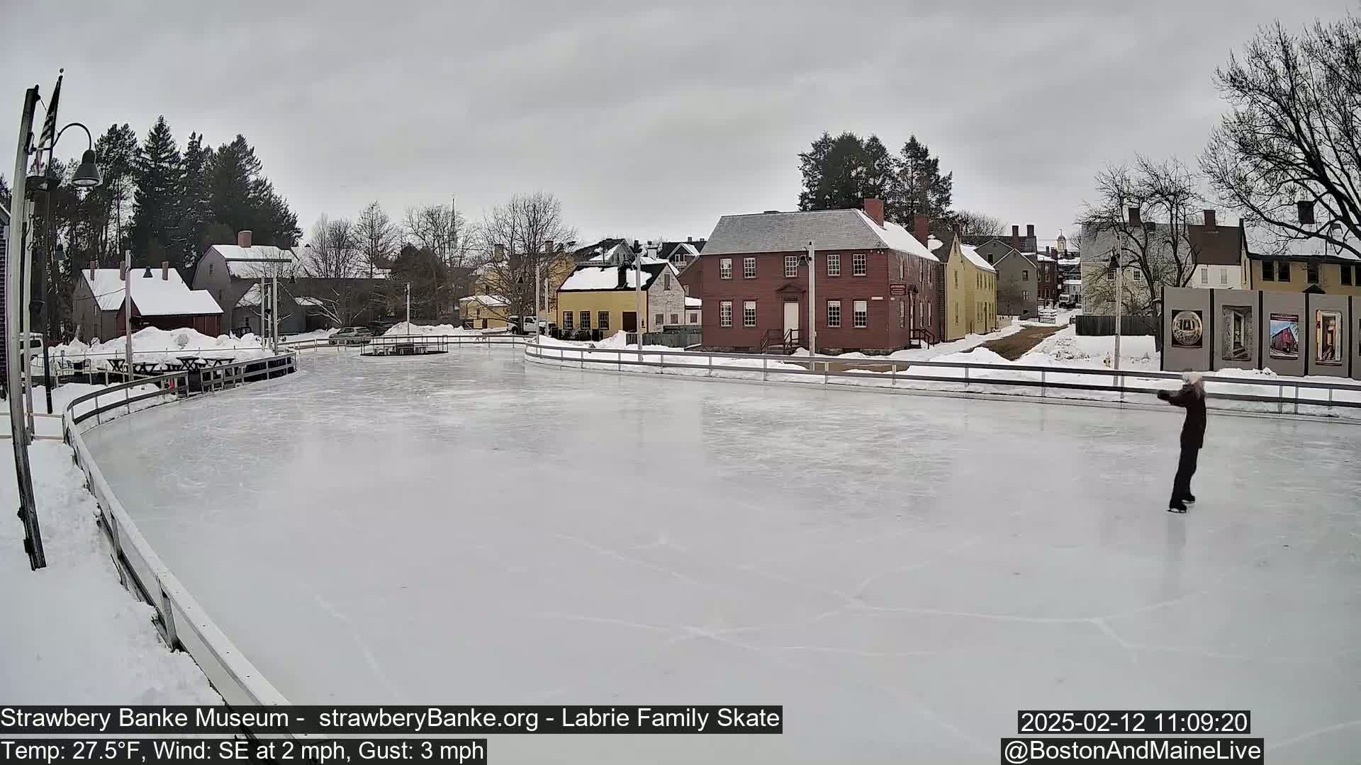 A person ice skates on a rink in front of historic buildings under an overcast sky.