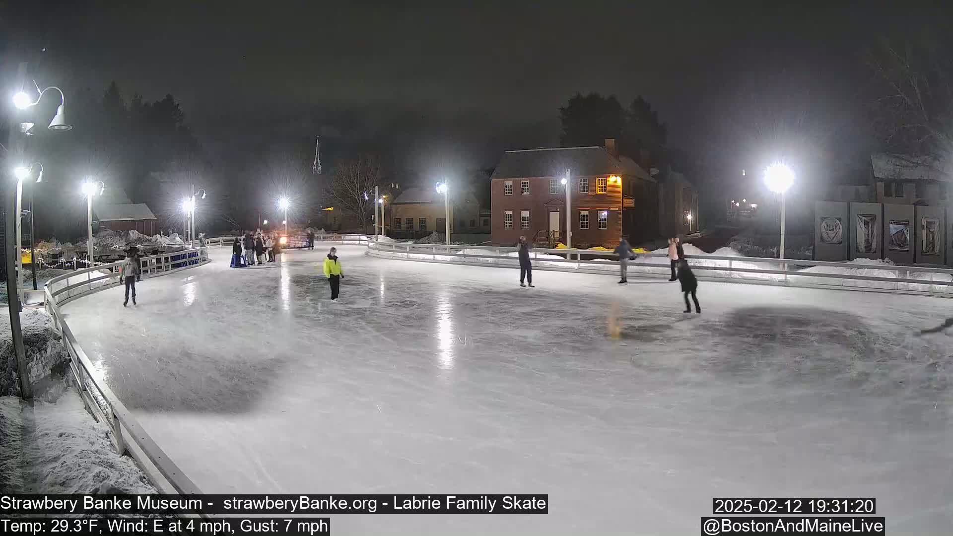 Several people ice skate on an outdoor rink at night, illuminated by lights, with a light wind.