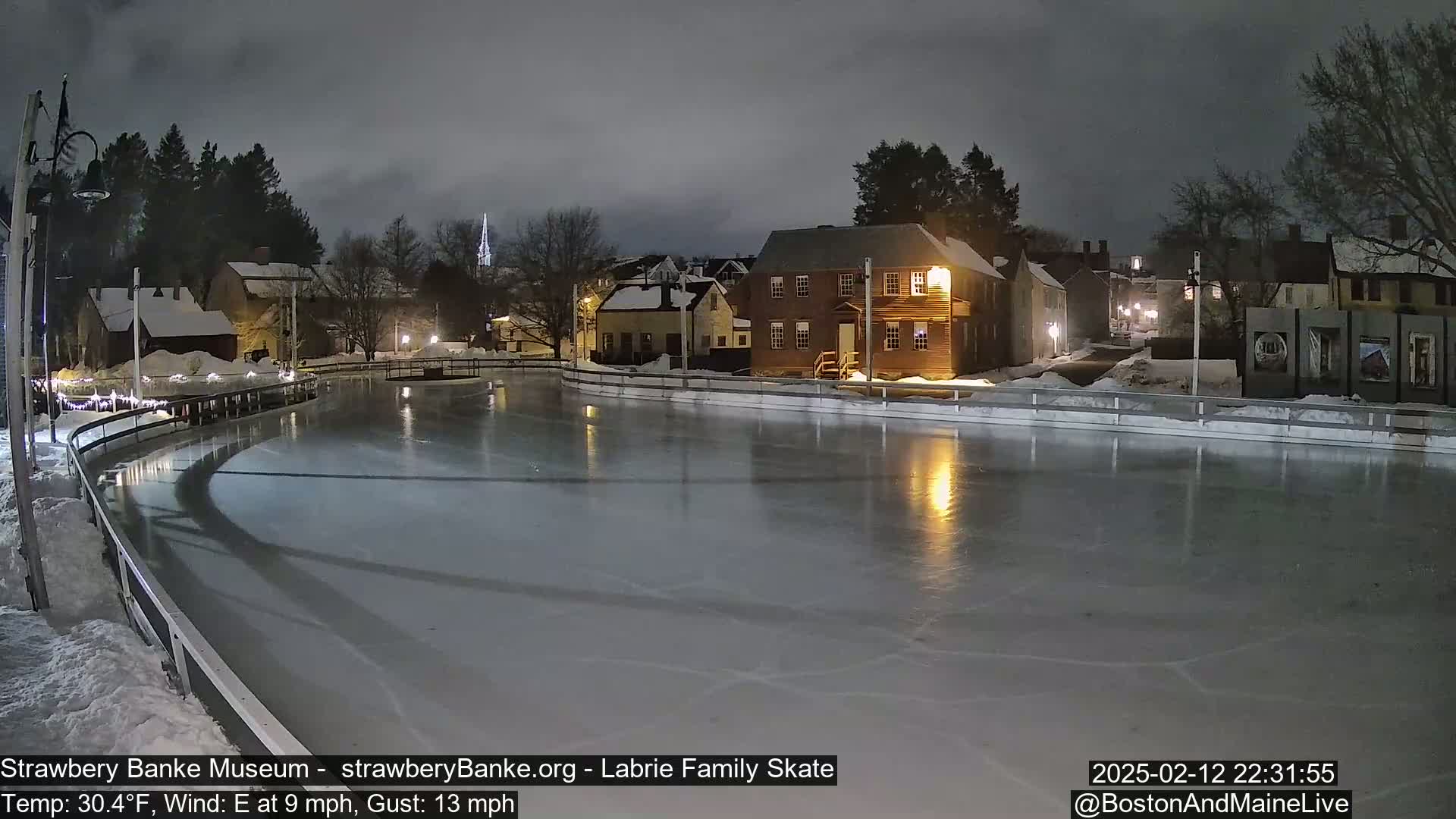 A nighttime view of an outdoor ice skating rink illuminated by lights, surrounded by snow-covered buildings and trees under a cloudy sky.