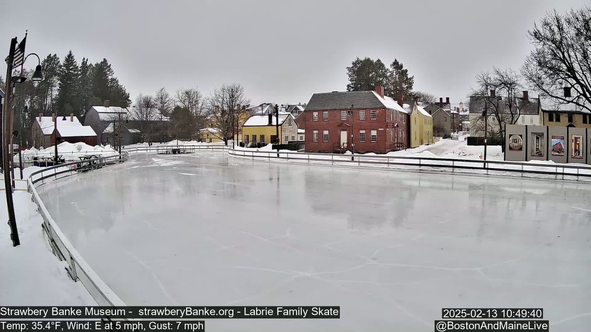 A frozen ice skating rink sits in front of several historic-looking buildings on an overcast day with an east wind.