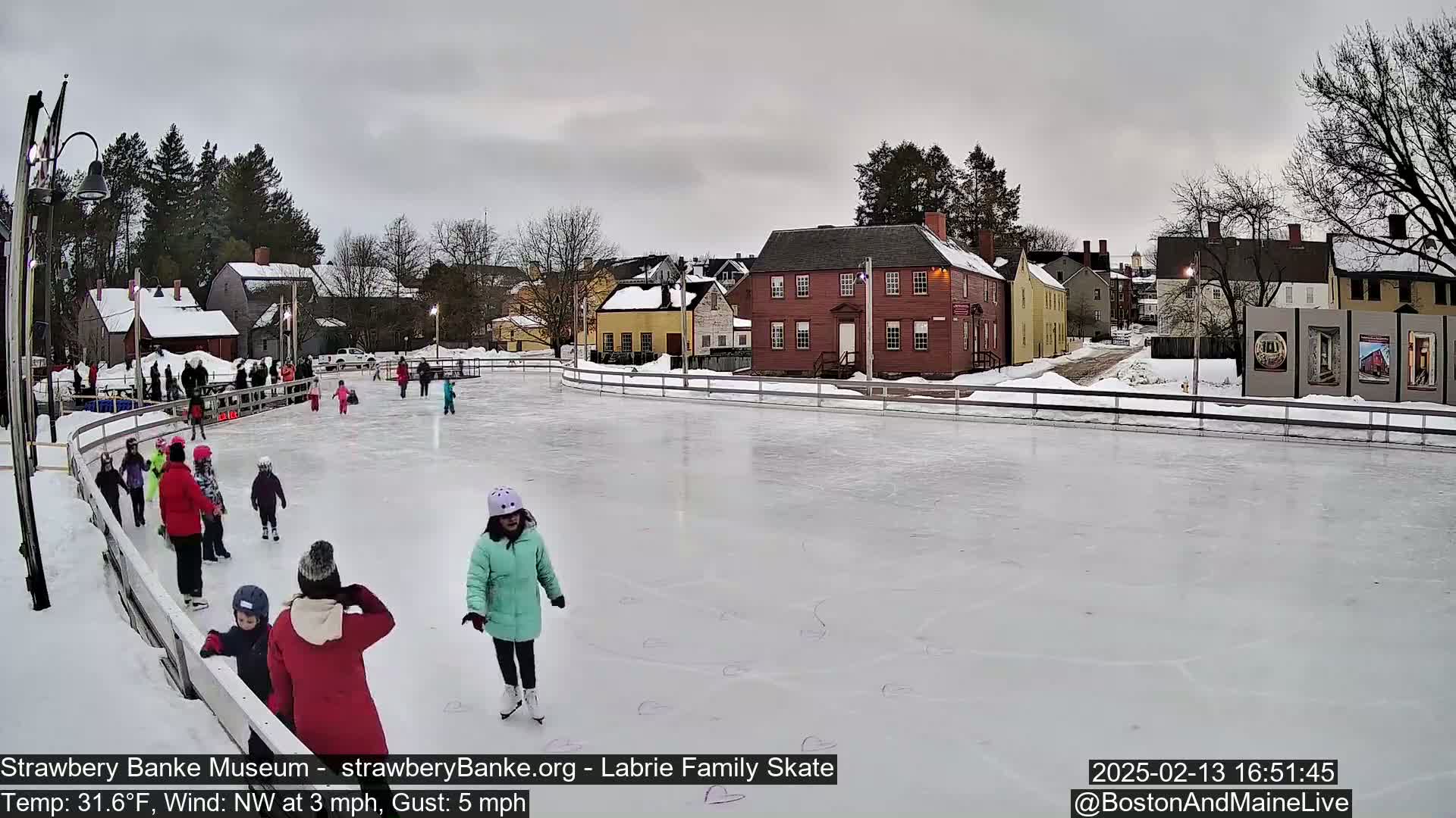 Children ice skate on an outdoor rink next to historic buildings under an overcast sky.