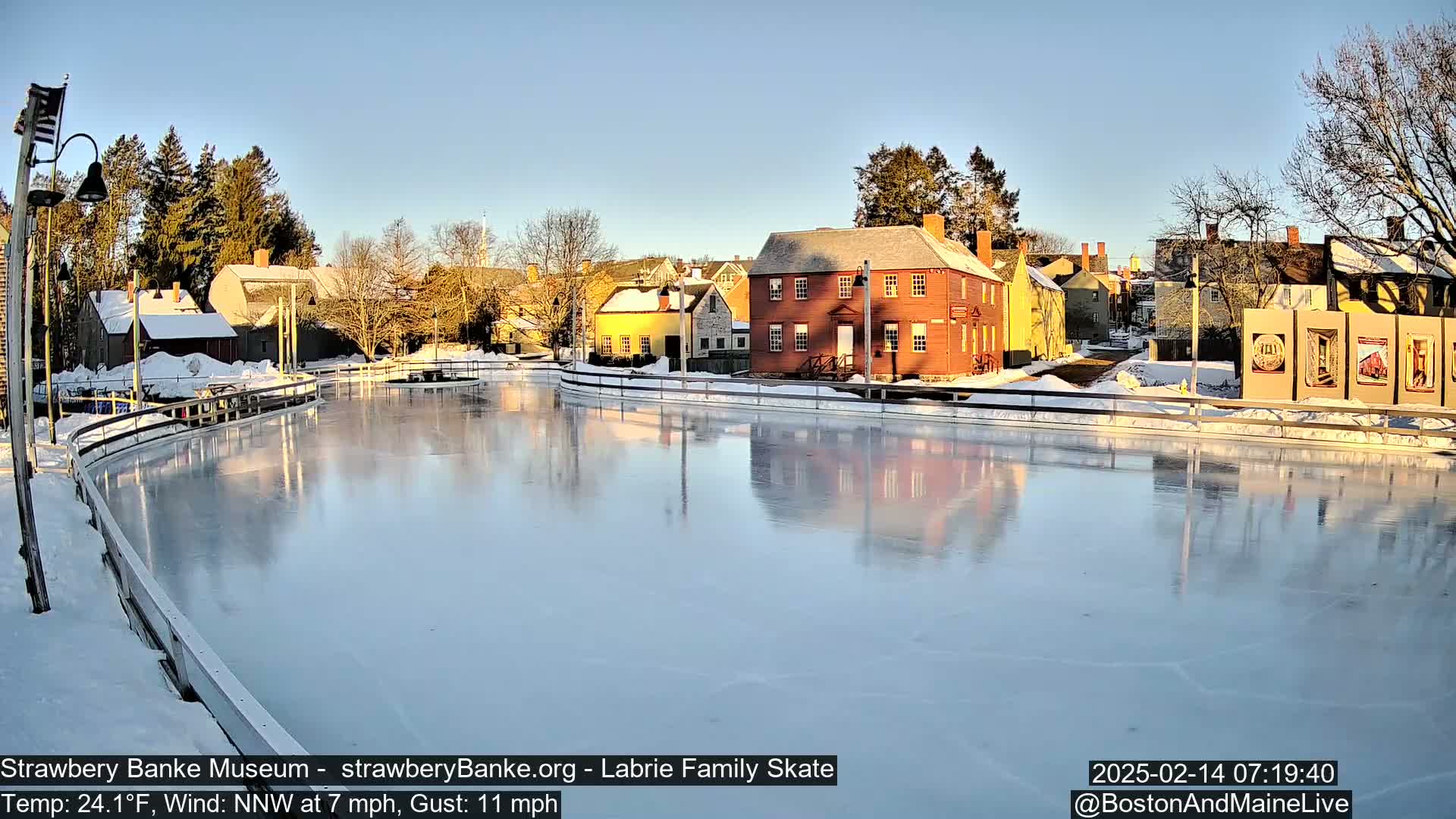 A smooth, icy skating rink reflects the surrounding colonial-style buildings under a clear, sunny sky in a snowy winter setting.
