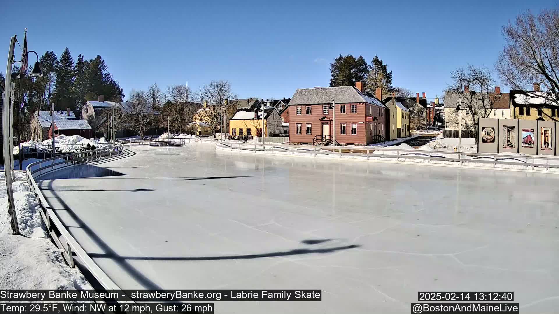A frozen outdoor skating rink is situated in front of several historic-looking buildings on a sunny day with a northwest wind.