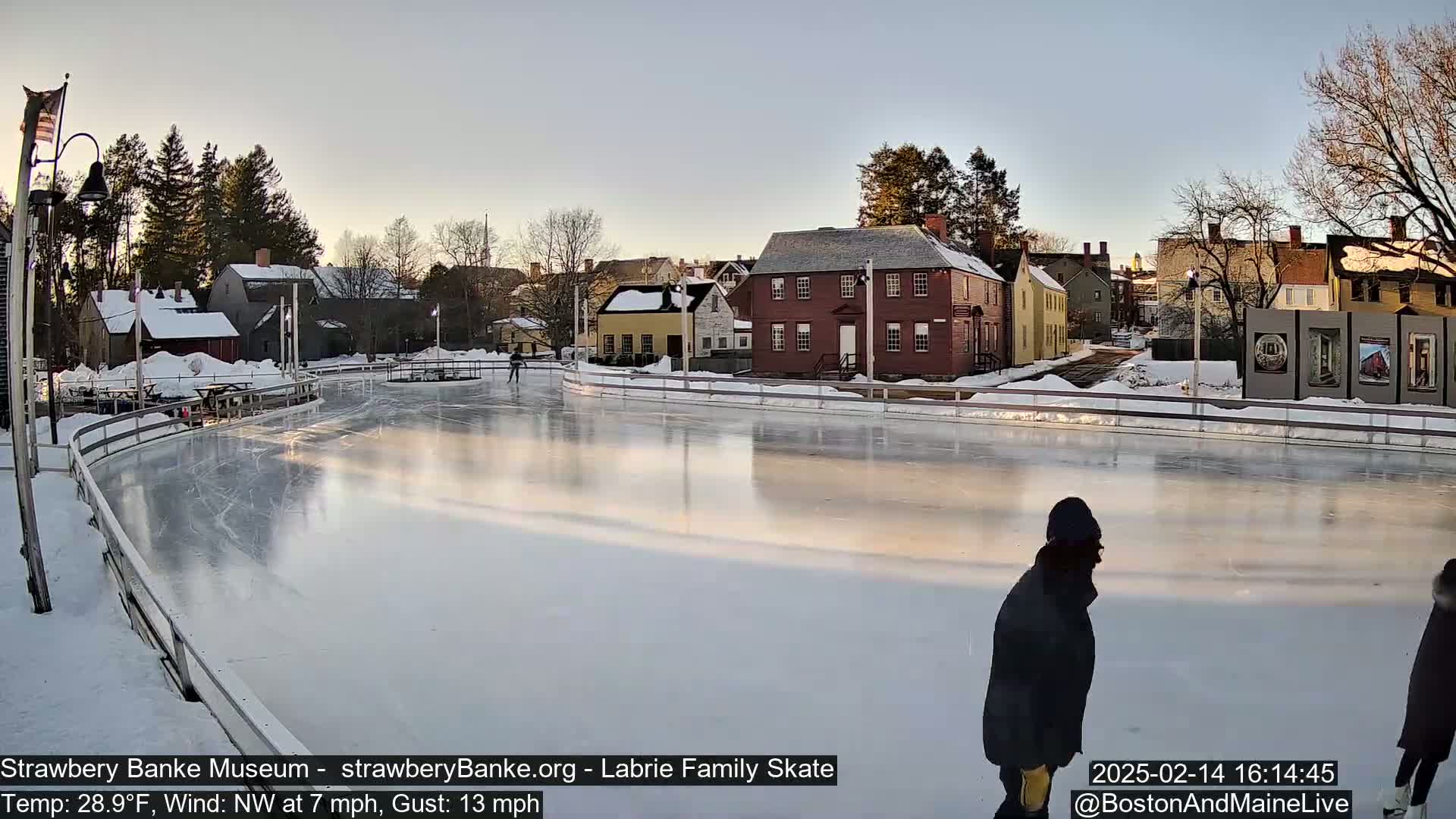 An outdoor ice skating rink, surrounded by snow-covered buildings and trees, is mostly empty except for a few skaters under a clear, bright sky.