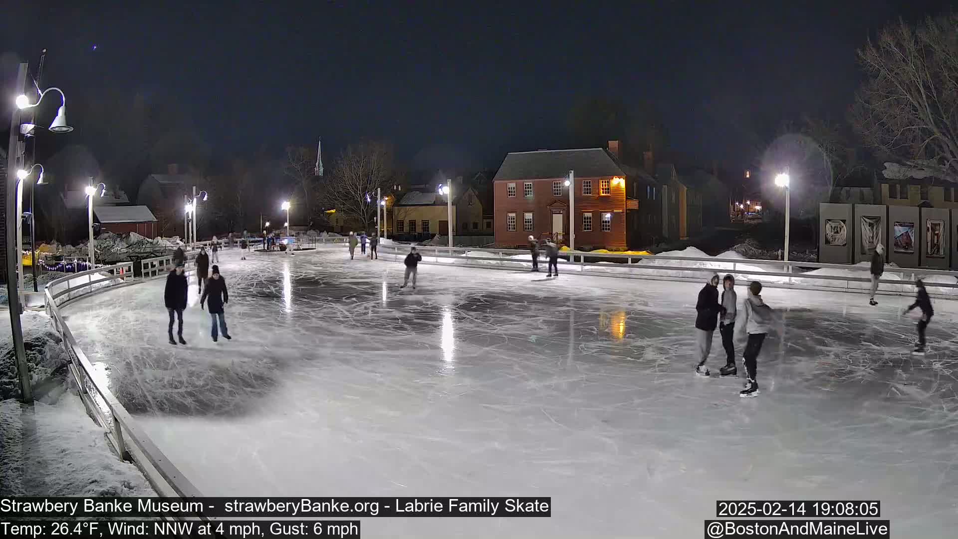 Several people ice skate at night on an outdoor rink illuminated by lights, under a clear, dark sky.
