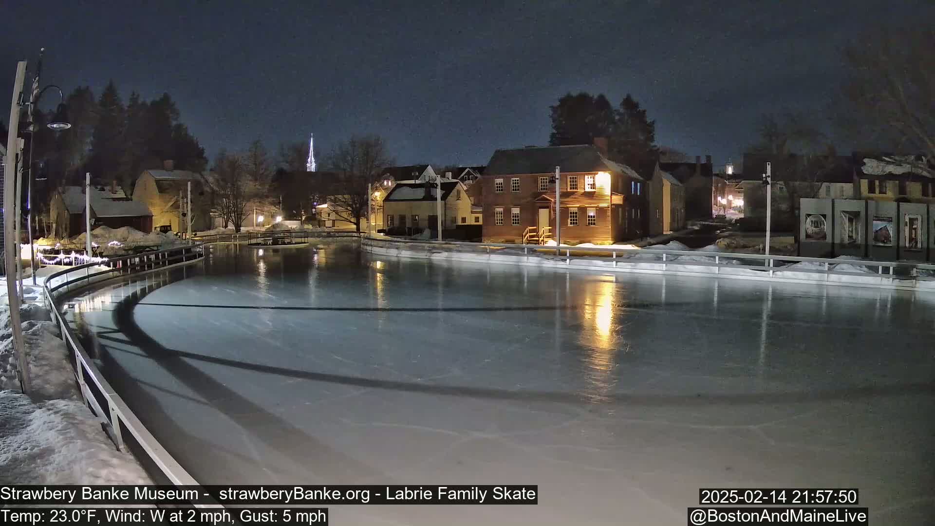 A nighttime view of an outdoor ice skating rink, illuminated, with historic buildings and a church spire in the background under a clear, dark sky.