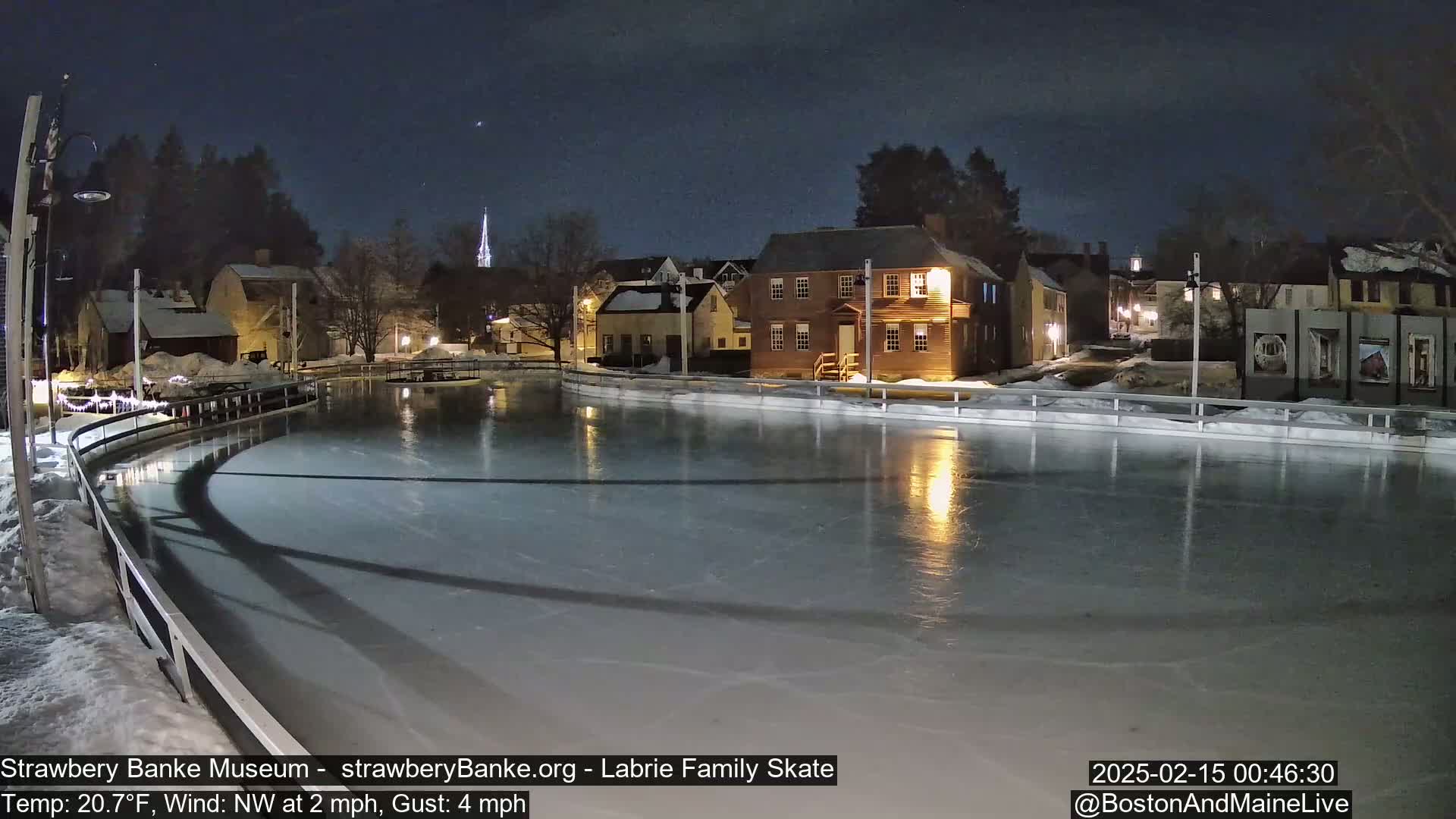 A nighttime view of an ice skating rink, illuminated, next to historic buildings under a clear, dark sky.