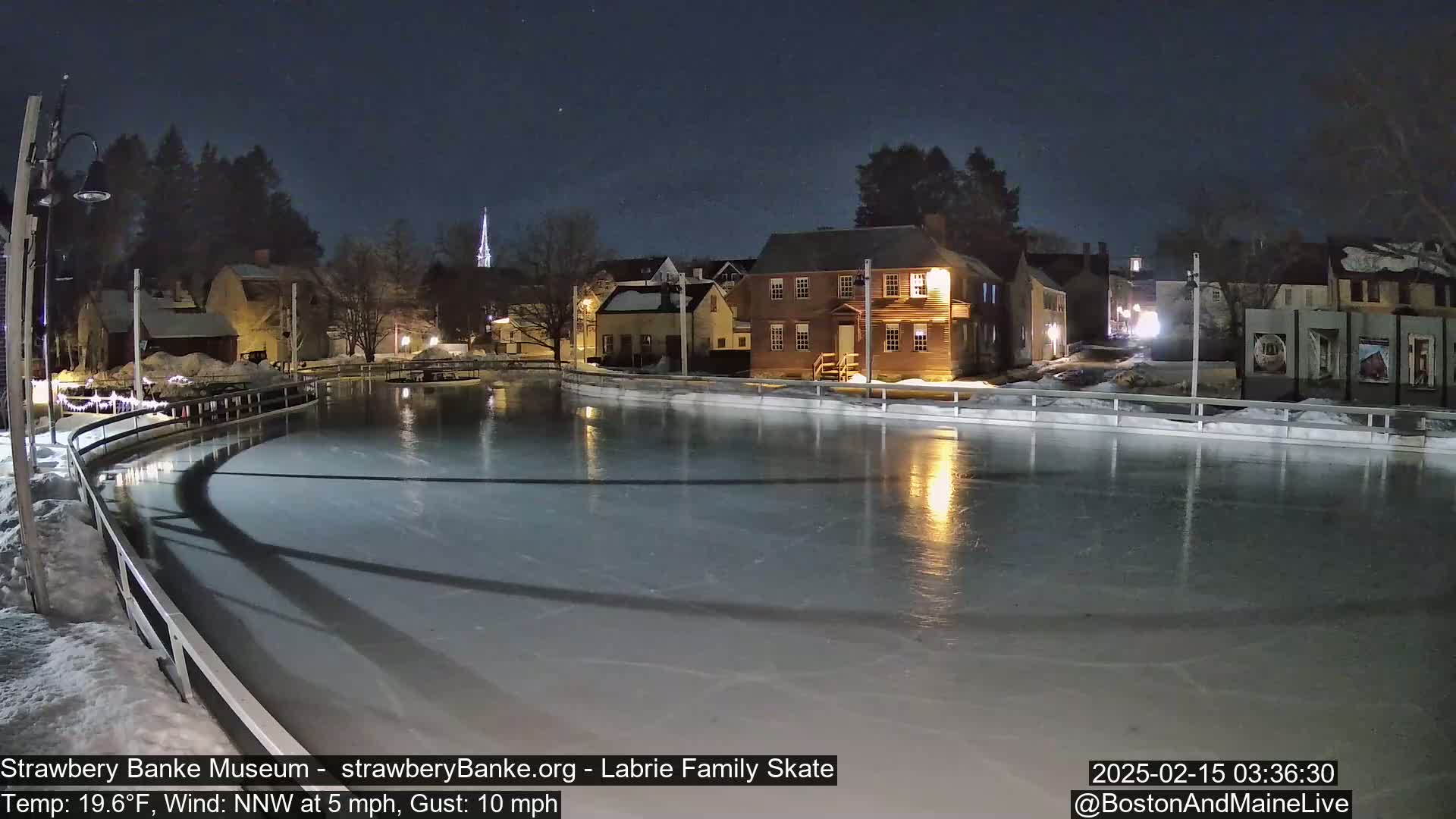 A nighttime view of an ice skating rink illuminated by lights, surrounded by snow-covered buildings under a clear, starry sky.