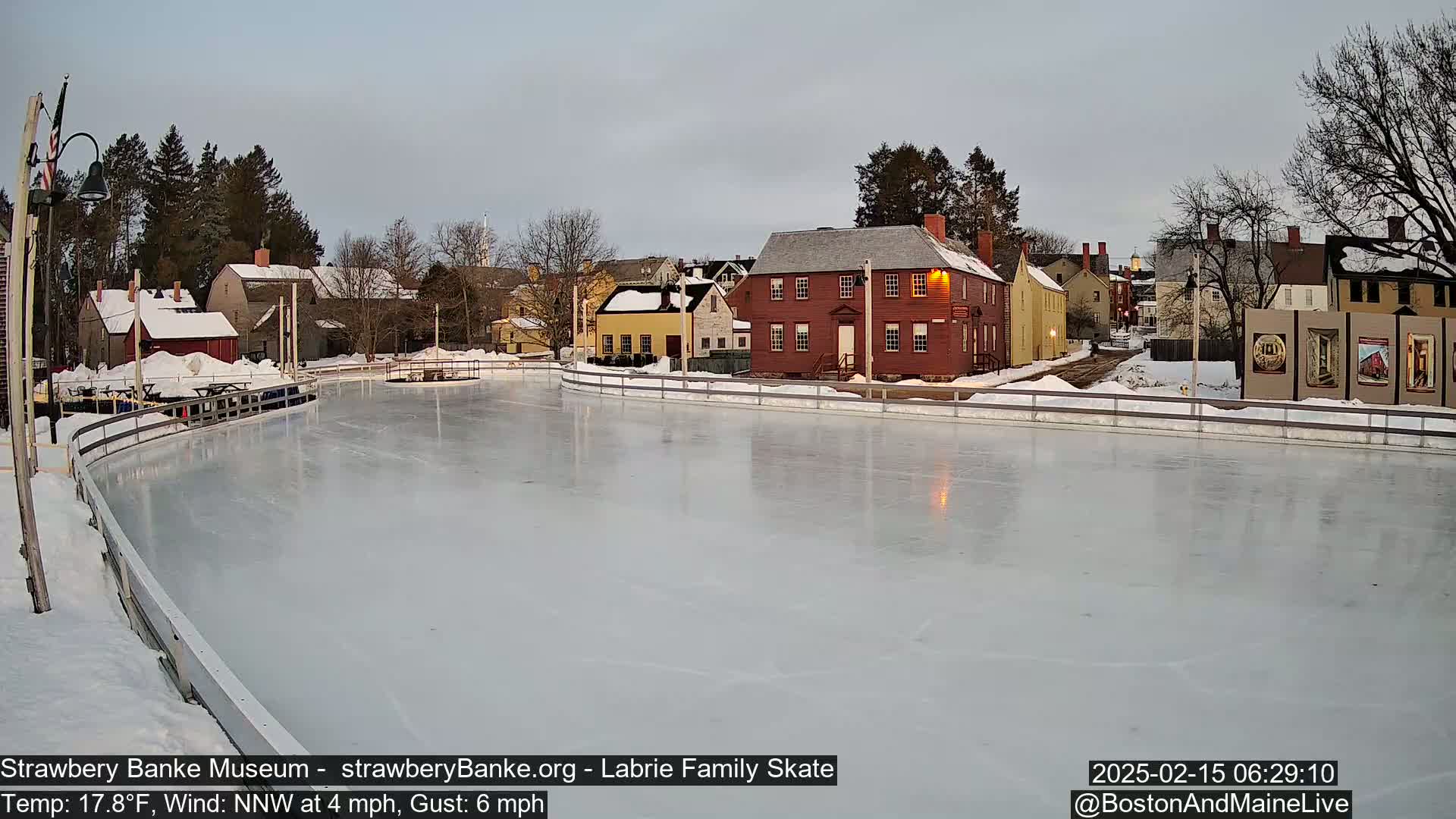 A frozen ice skating rink sits in front of a row of historic-looking buildings on a partly cloudy, cold morning.