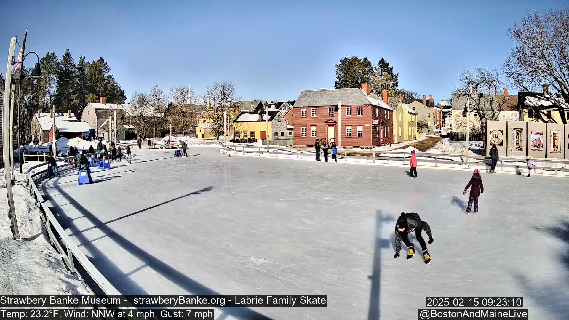 An outdoor ice-skating rink, surrounded by historic buildings under a clear blue sky, features several people ice skating on a sunny, cold day.