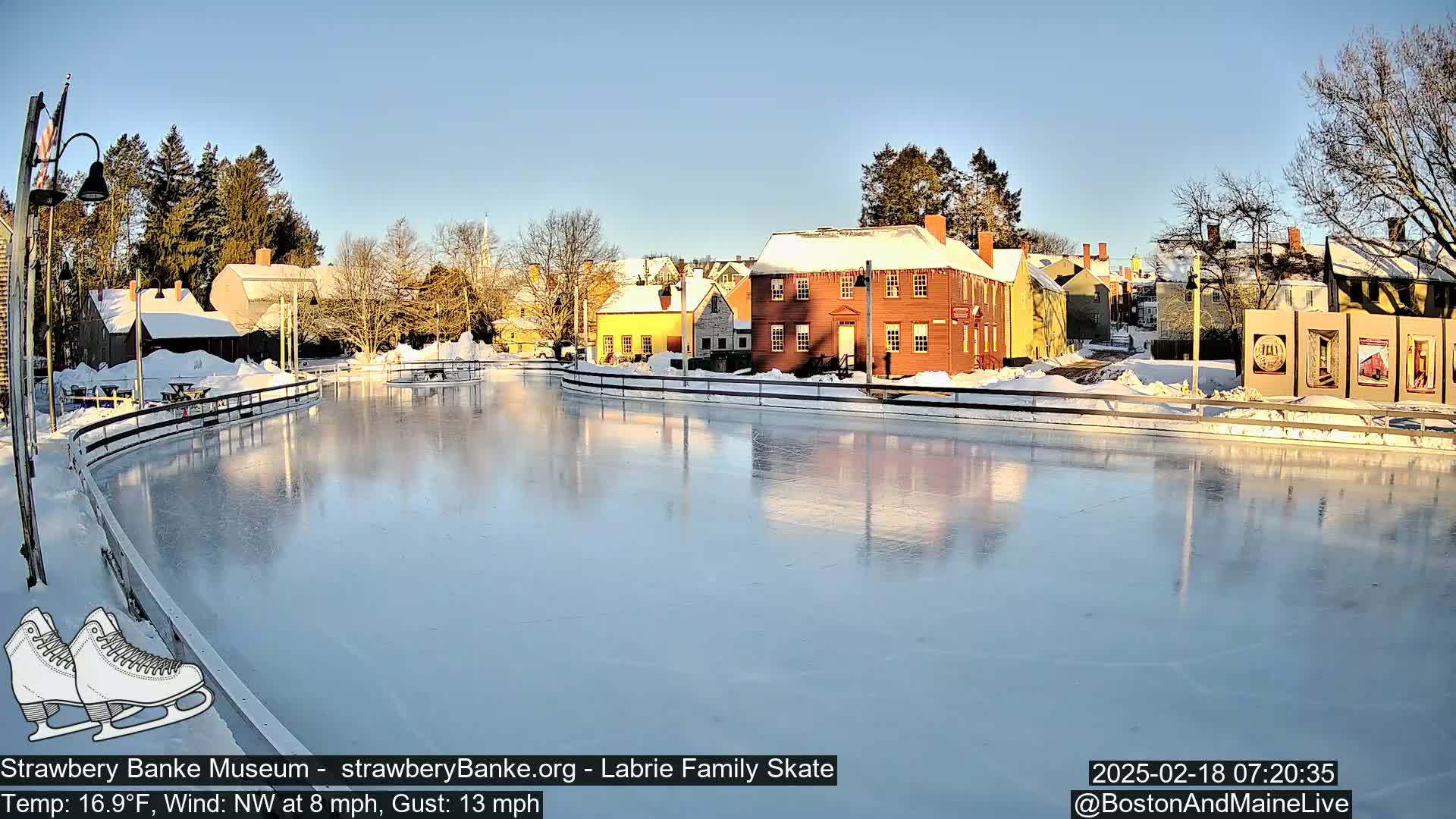A frozen skating rink reflects the sunlit, snow-covered buildings of a village under a clear, blue sky with a northwest wind.