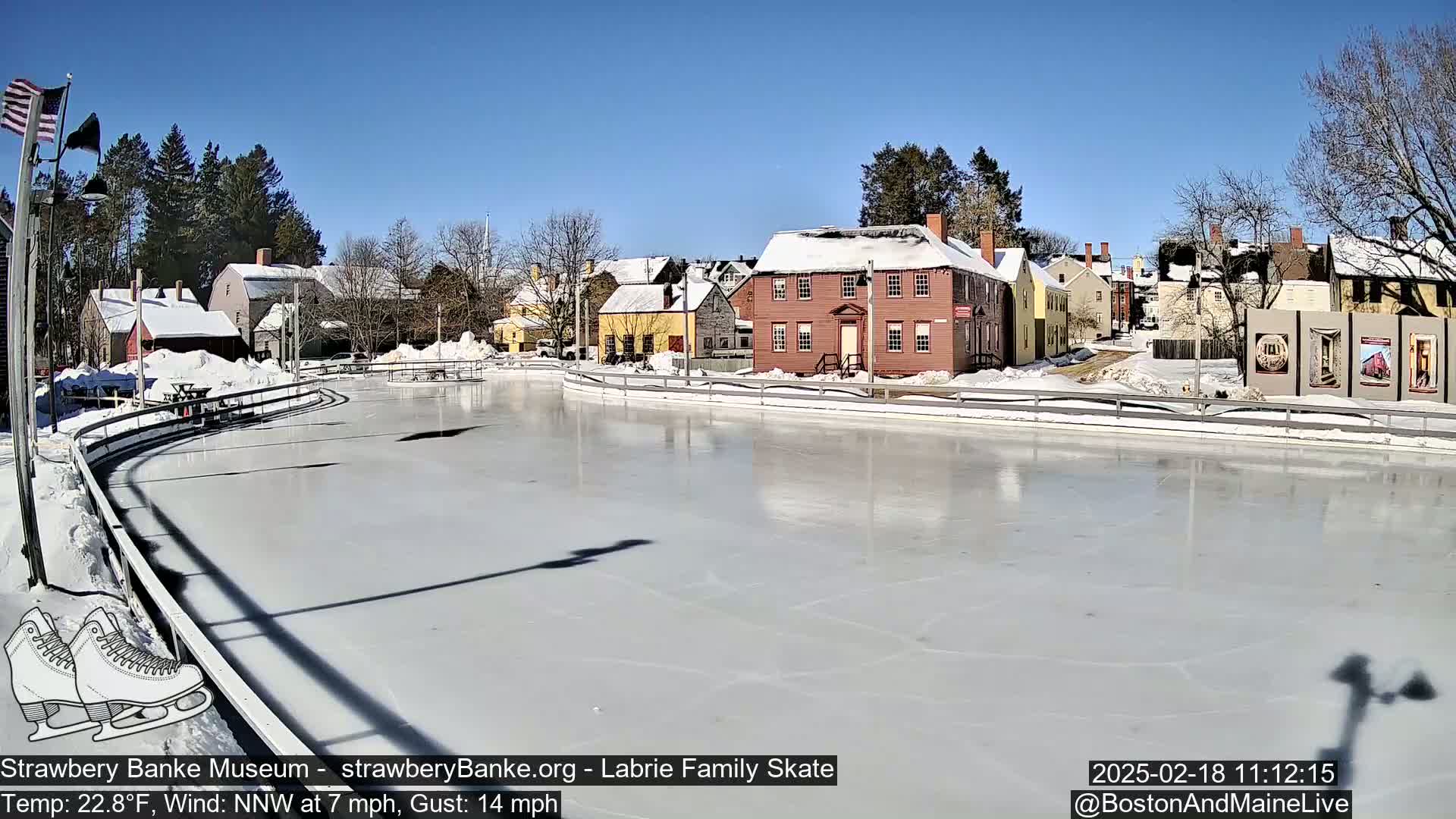 A frozen outdoor skating rink sits in front of a row of snow-covered colonial-style buildings under a clear blue sky.