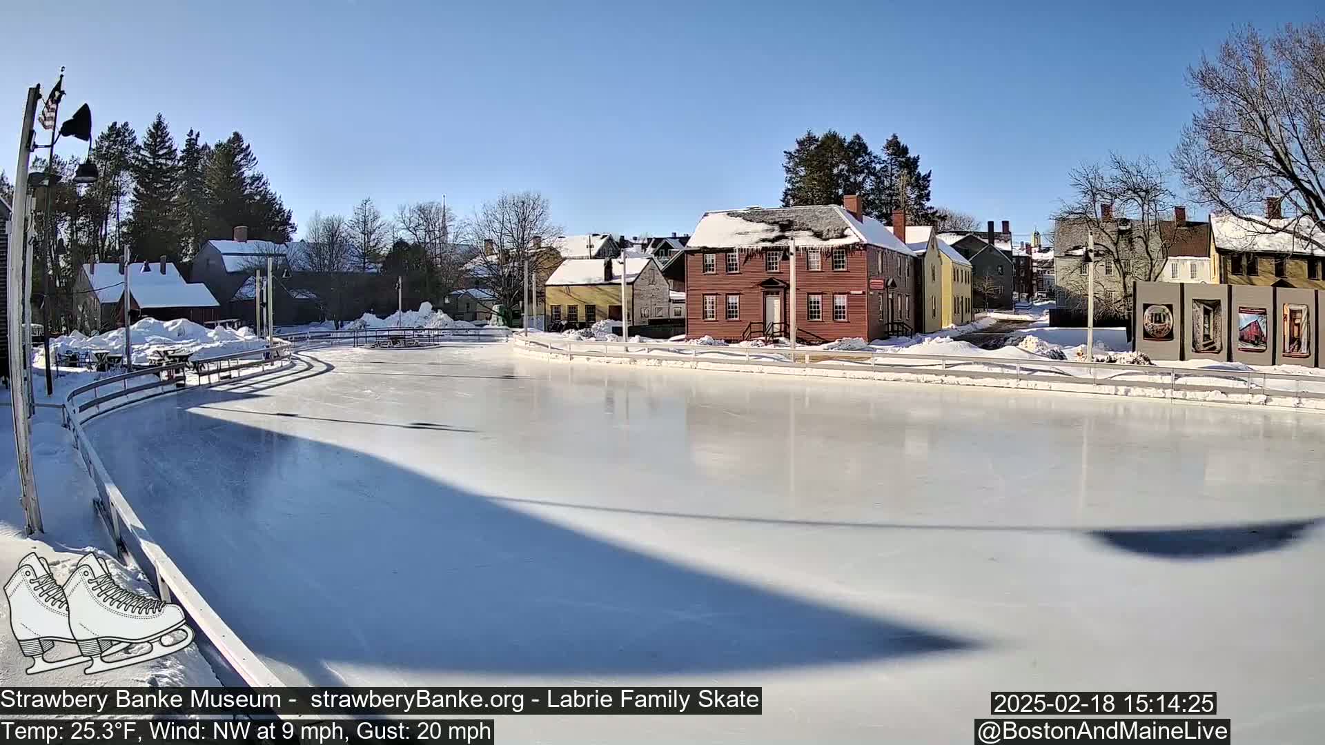 A sunny, snow-covered outdoor ice rink is situated beside several historic-looking buildings under a clear blue sky.