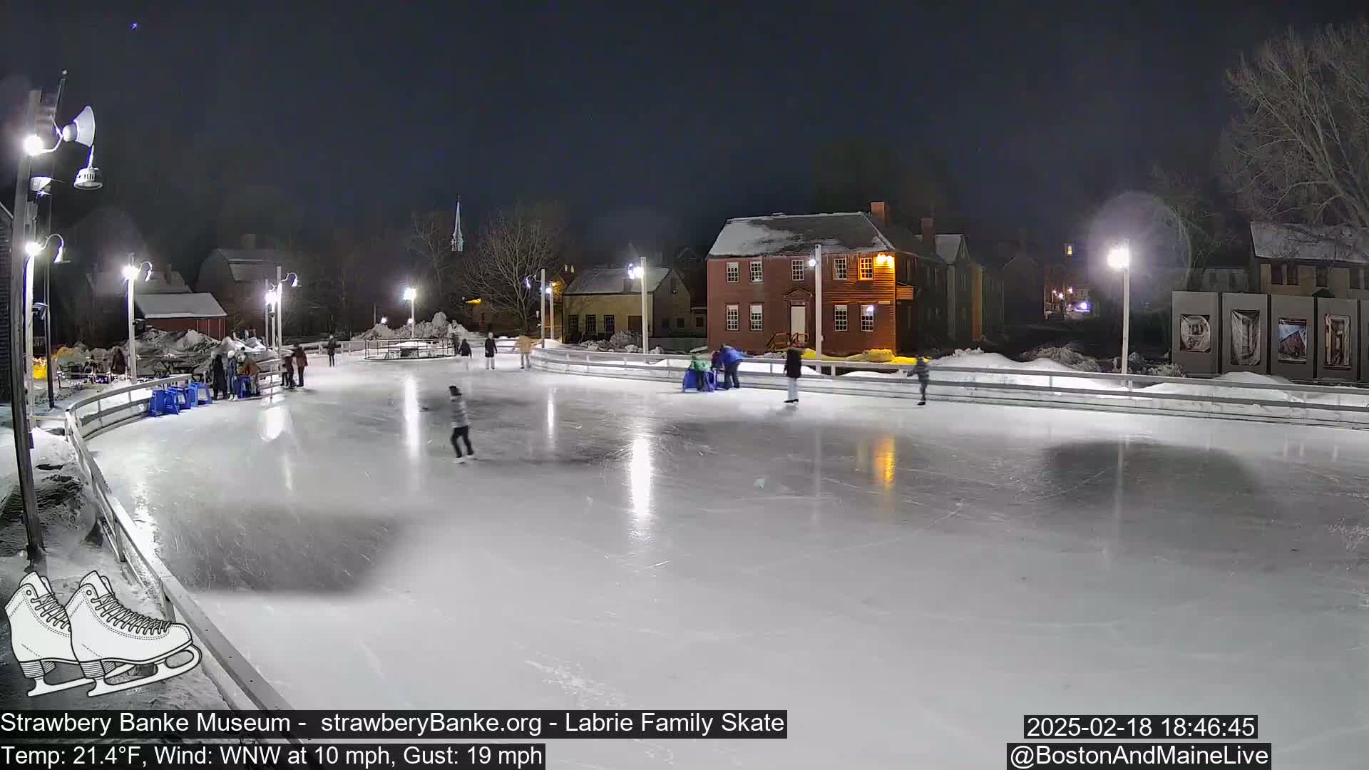 At night, under a clear sky, several people ice skate on an outdoor rink illuminated by lights, with buildings visible in the background.