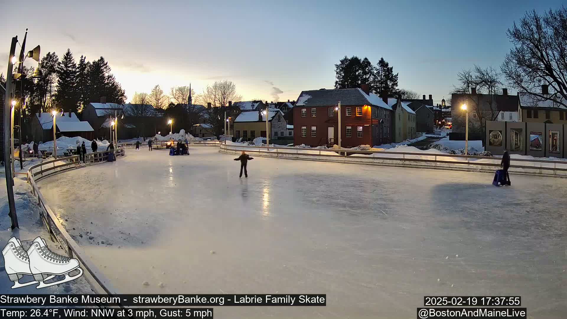 An outdoor ice skating rink at dusk, with several people ice skating, is surrounded by snow-covered buildings and trees under a twilight sky.