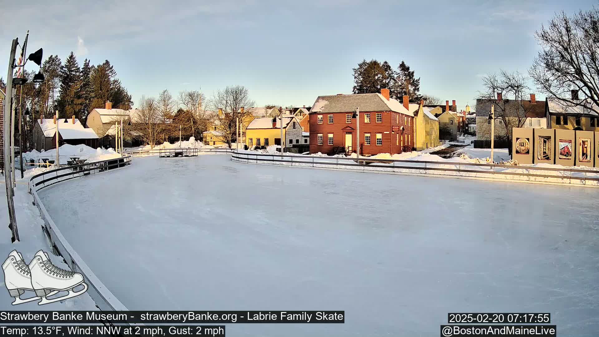 A snow-covered outdoor ice rink is situated in front of several historic-looking buildings under a mostly clear sky.