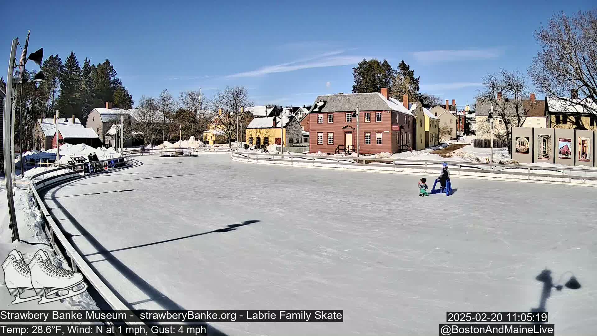 A sunny outdoor ice-skating rink, surrounded by snow-covered historical buildings, with two children skating.